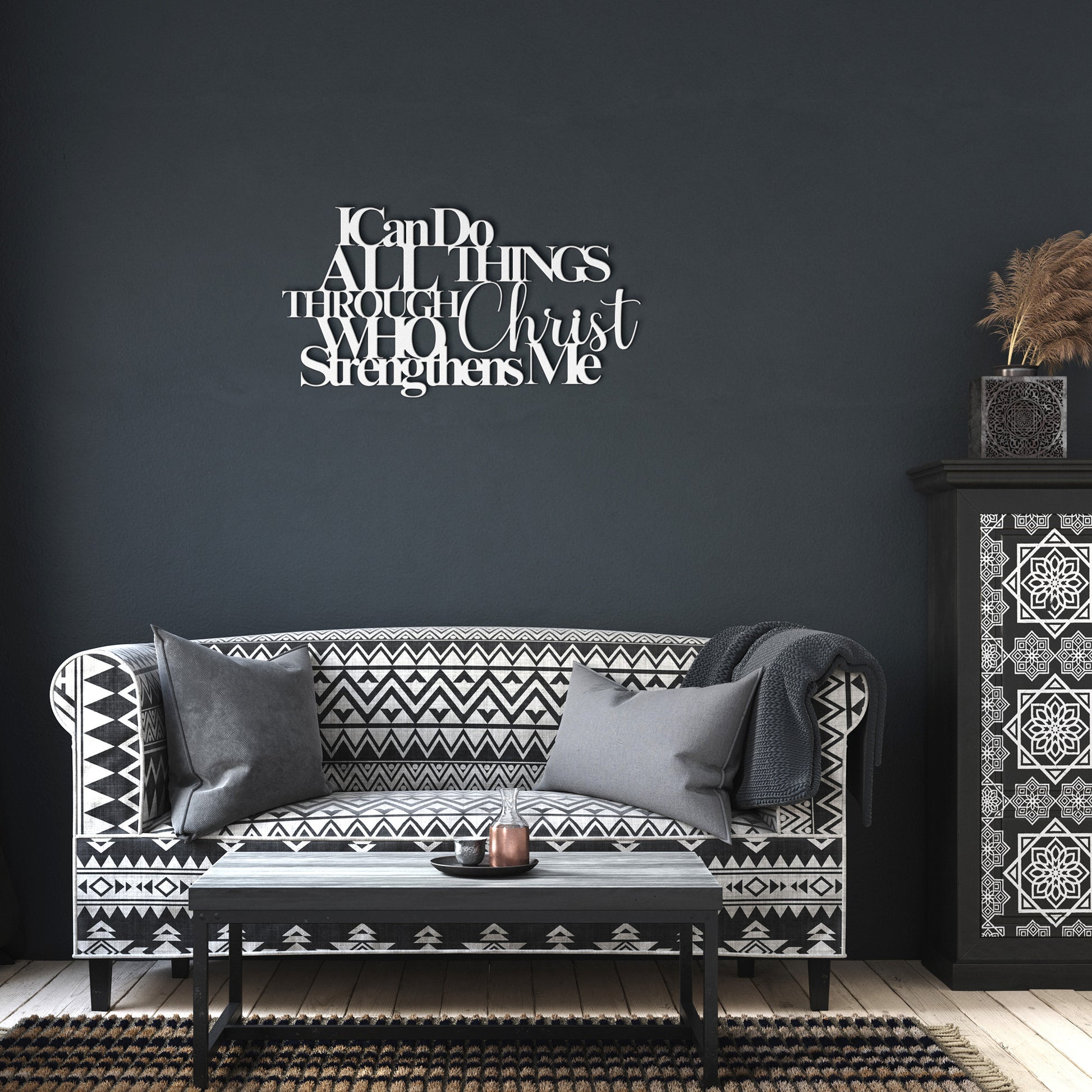 A living room features a patterned black-and-white sofa with two gray cushions, a black coffee table, a decorative cabinet with patterned designs, and the "I Can Do All Things Through Christ Who Strengthens Me Christian Metal Sign" on the wall.