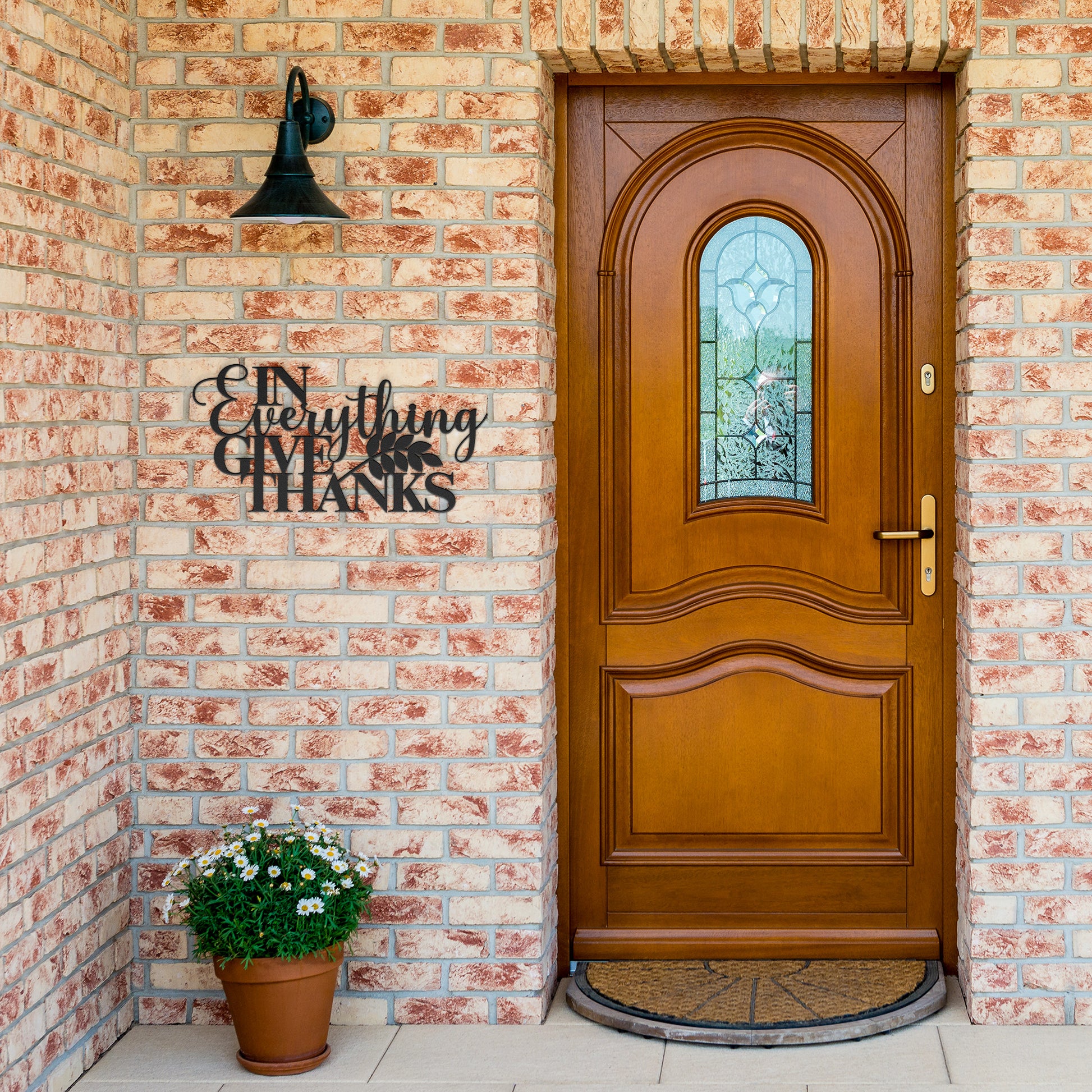 A wooden door with an arched glass panel set in a brick wall features a potted plant sitting next to it. A powder-coated metal sign displaying the message "In Everything Give Thanks Christian Metal Art" hangs beside the door.