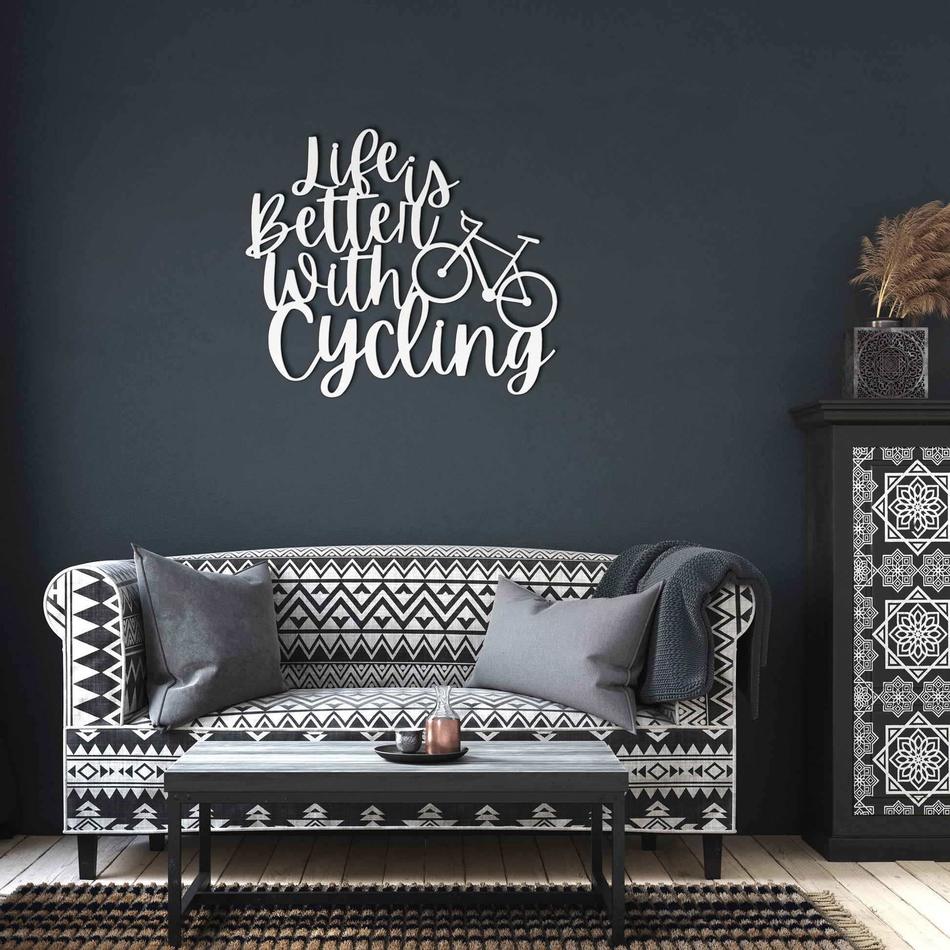 A brick wall with a wooden door, a potted plant, a black outdoor light, and the "Life is Better with Cycling Metal Sign - Road Bike Edition," reading "Life's Better with Cycling" alongside a bicycle graphic, perfect for stylish home decor.