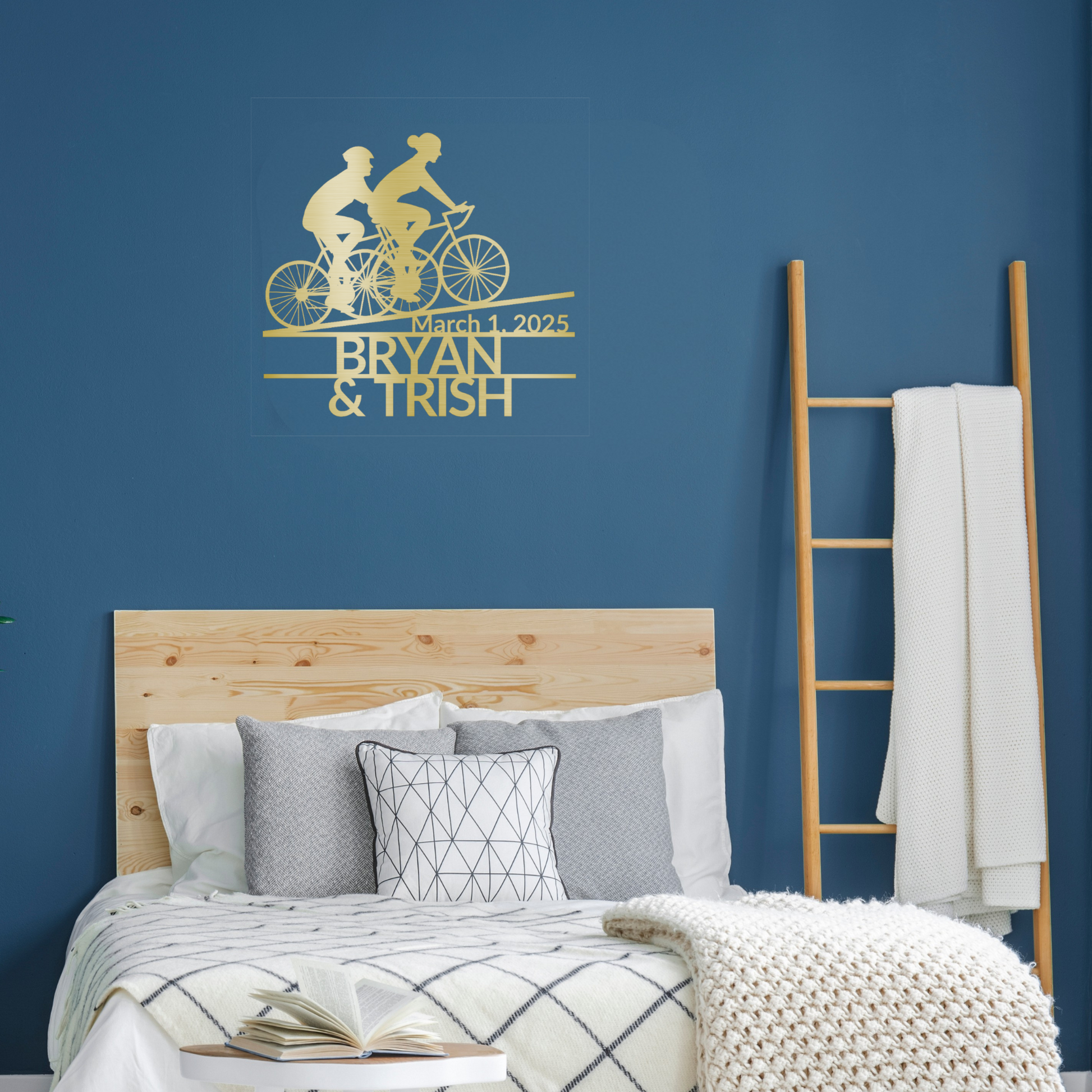 A modern bedroom features a wooden bed, gray and white bedding, a ladder with a blanket, and the Custom Cyclist Couple Metal Sign by MEM Design Studio personalized for Bryan & Trish with their date, March 1, 2025.