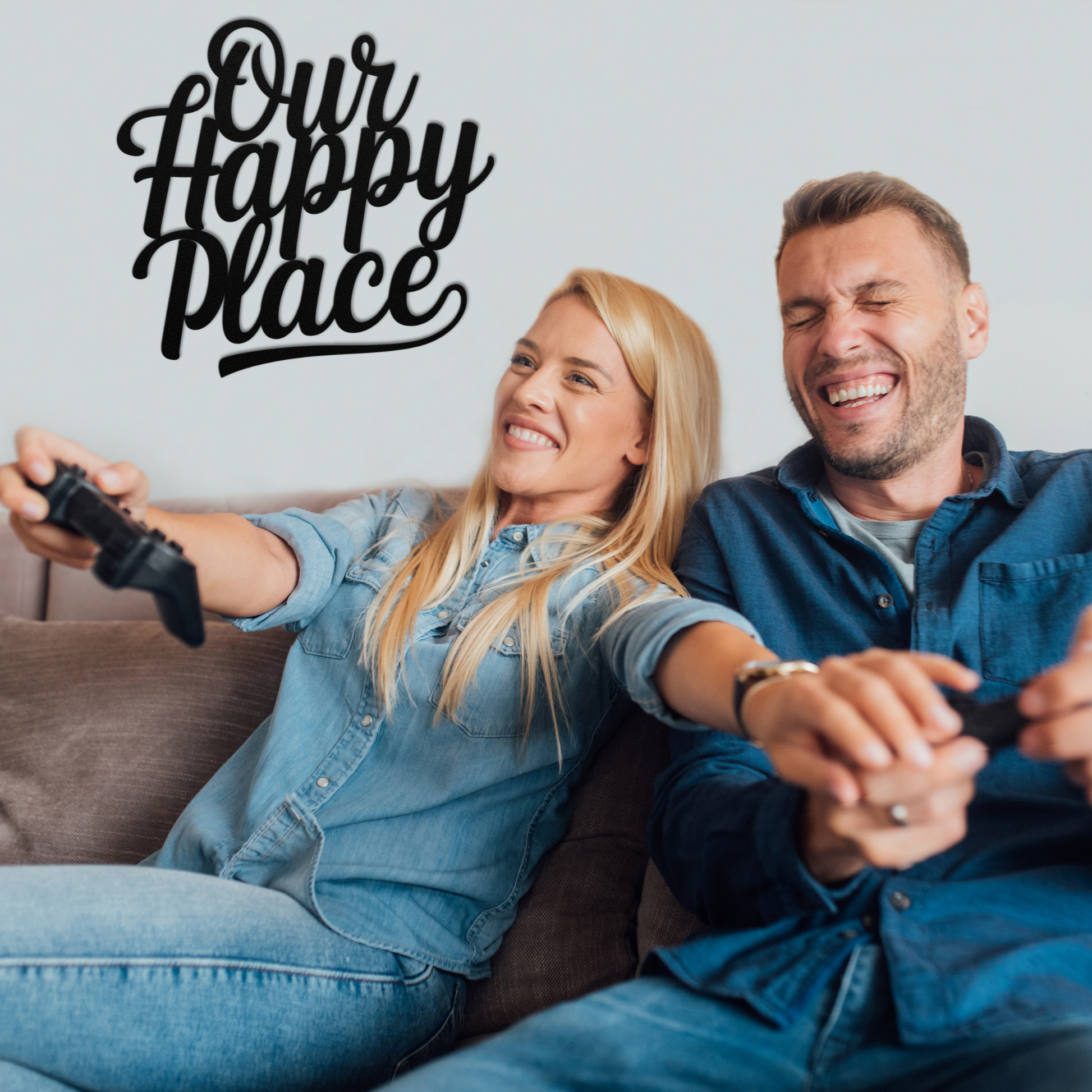 A smiling couple sits on a couch playing video games, both laughing in denim shirts. Above them, "Our Happy Place Metal Wall Art" from MEM Design Studio adds a personal, customizable touch to their living space.