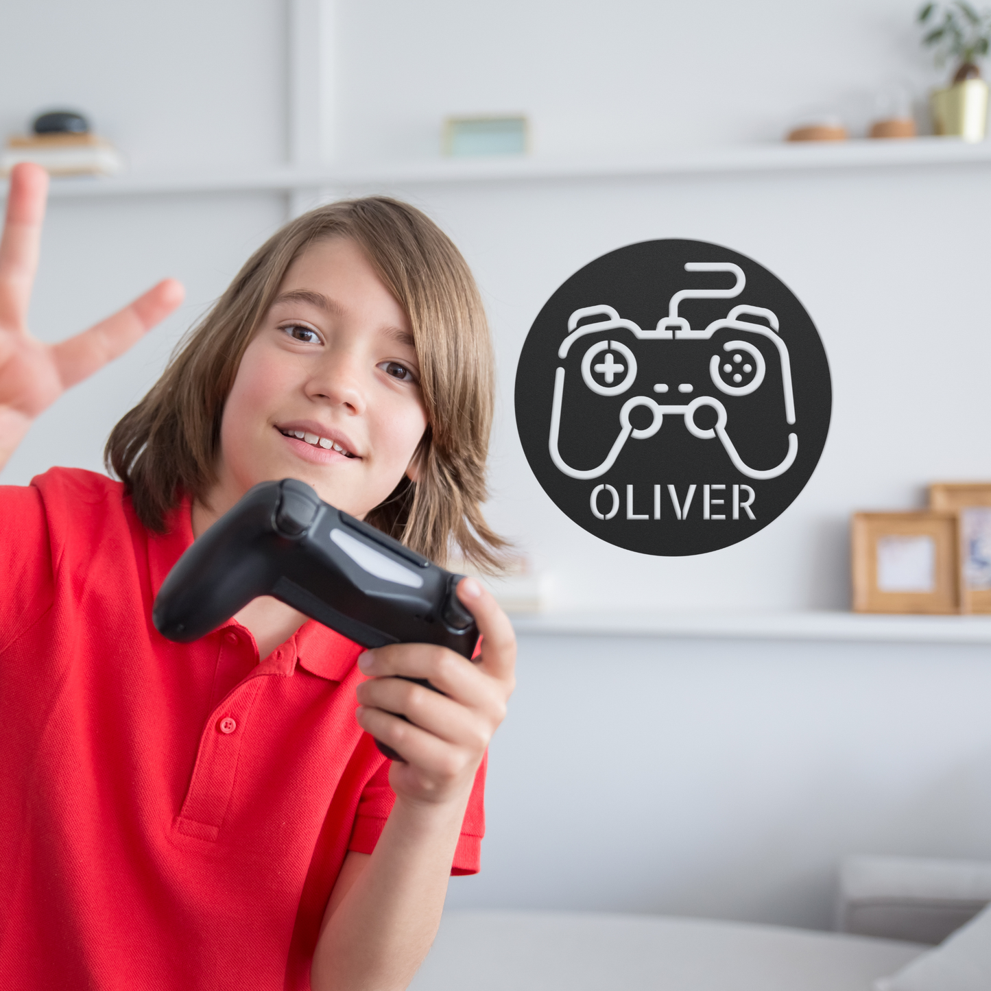 A smiling child in a red shirt holds a video game controller and flashes a peace sign. Behind, a Personalized Gamer Controller Metal Sign with "Oliver" decorates the wall, with shelves and decor visible in the background.