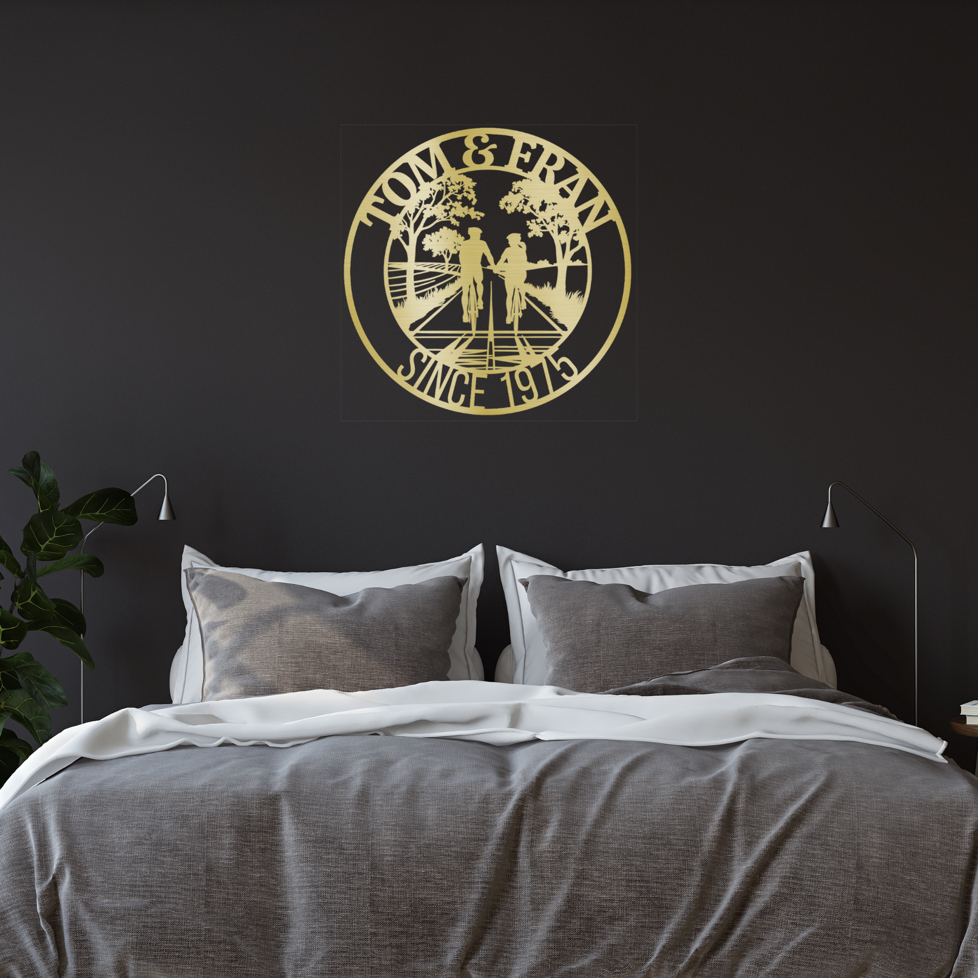 A cozy bedroom with a neatly made bed, gray bedding, two reading lamps, and the Personalized Cyclist Couple Metal Wall Art by MEM Design Studio above the headboard, customized with “TOM & FRAN SINCE 1975” and an outdoor scene.