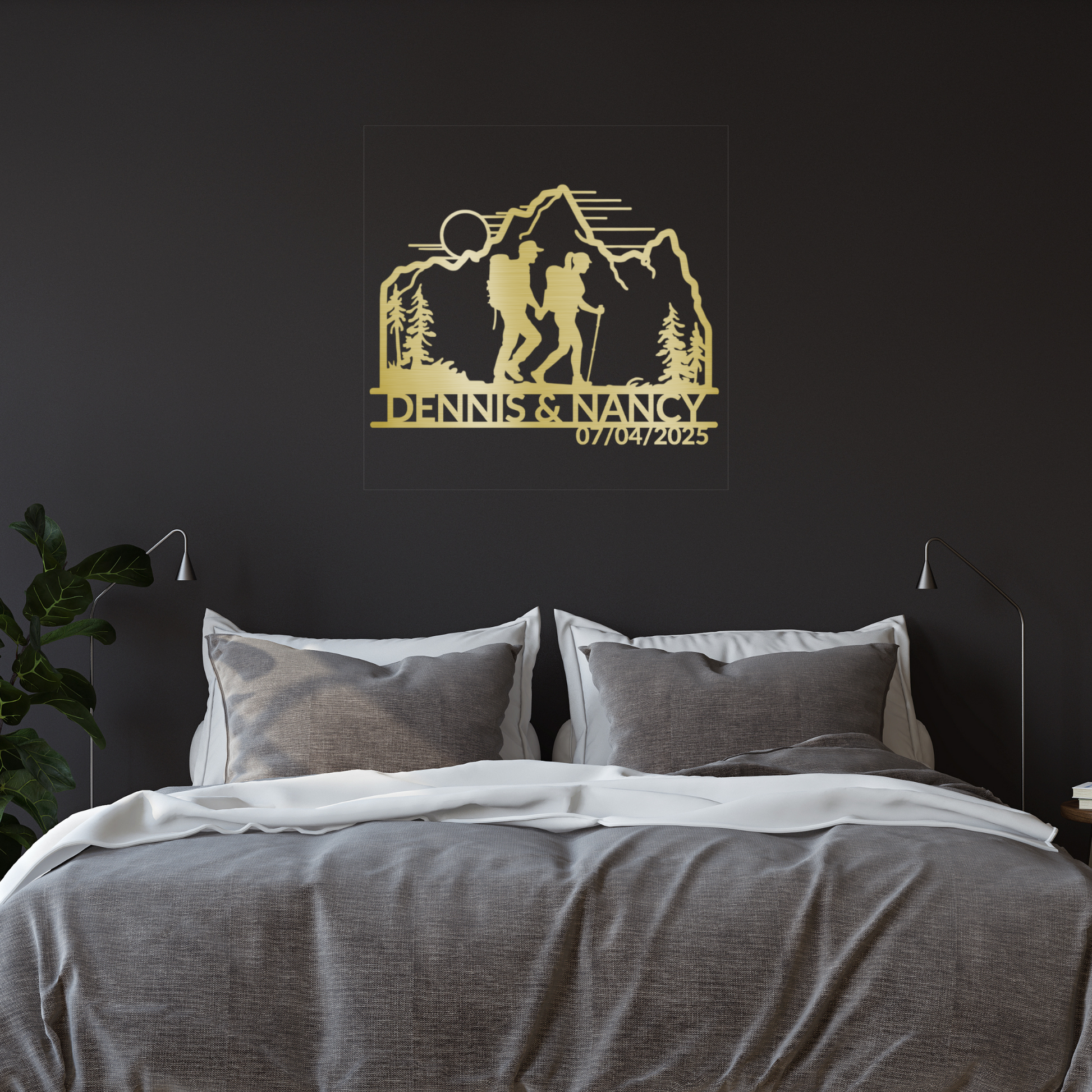 A modern bedroom showcases a double bed with gray bedding, white pillows, and the Personalized Hiking Couple Metal Wall Art above the bed, depicting a hiking couple, mountains, trees, and "DENNIS & NANCY" with the date 07/04/2025.