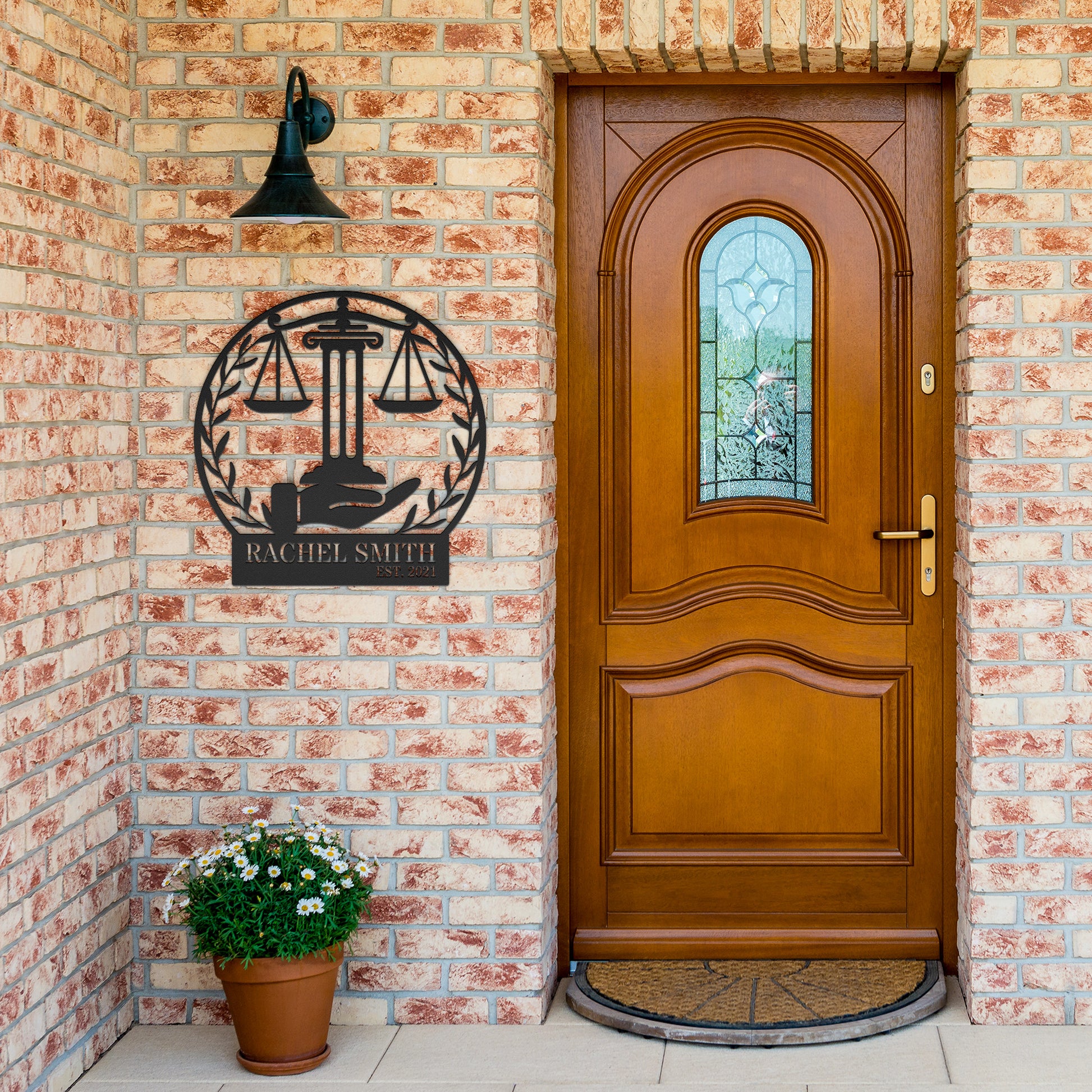 A brick exterior wall with a wooden door featuring an arched window. Beside the door, a black metal sign with a "Scales of Justice" design reads "Rachel Smith." A potted plant with white flowers sits next to the door, adding a touch of home decor.