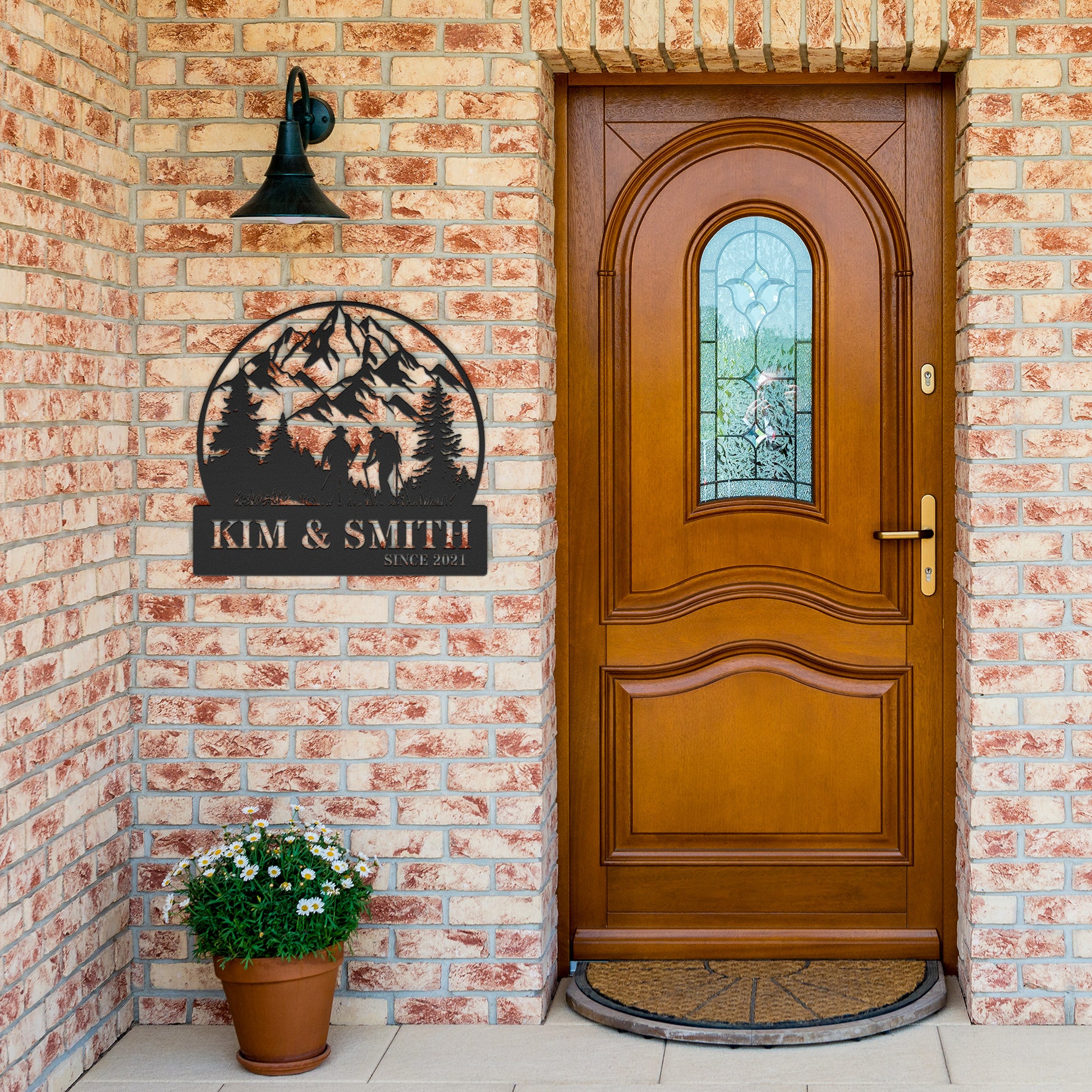 A brick house exterior with a wooden door, arched stained glass, wall-mounted lamp, potted flowers, and an 18 gauge steel decor sign reading "Kim & Smith," featuring the Updated Hiking Couple Metal Art with a mountain and tree silhouette that serves as a timeless piece of home decor.