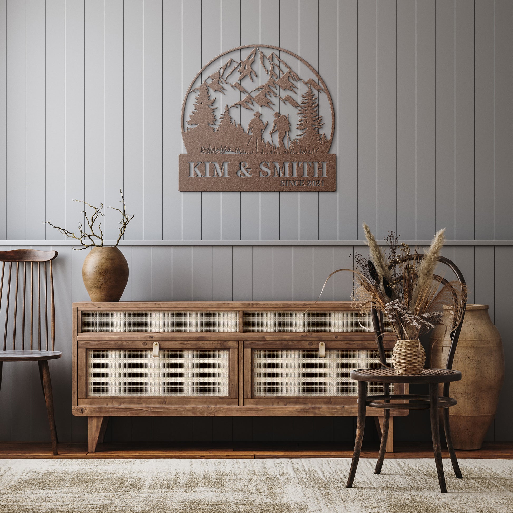 A brick house exterior with a wooden door, arched stained glass, wall-mounted lamp, potted flowers, and an 18 gauge steel decor sign reading "Kim & Smith," featuring the Updated Hiking Couple Metal Art with a mountain and tree silhouette that serves as a timeless piece of home decor.