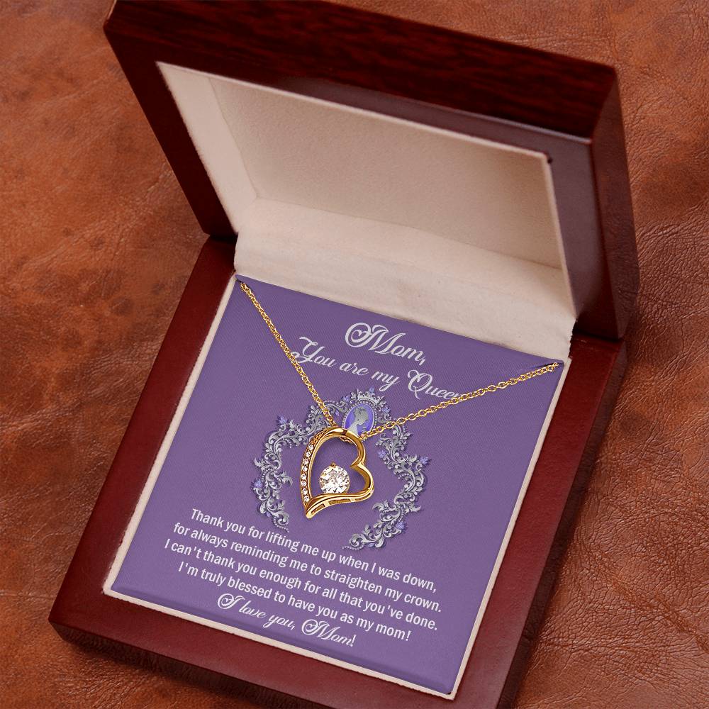 The "To Mom, Belongs To Me - Forever Love Necklace" is a gold heart-shaped necklace with cubic zirconia gemstones, elegantly displayed in an open wooden box lined with white fabric. It comes with a heartfelt message reading "Mom, You are my Queen" printed on a purple card inside, making it the perfect personalized gift for her.