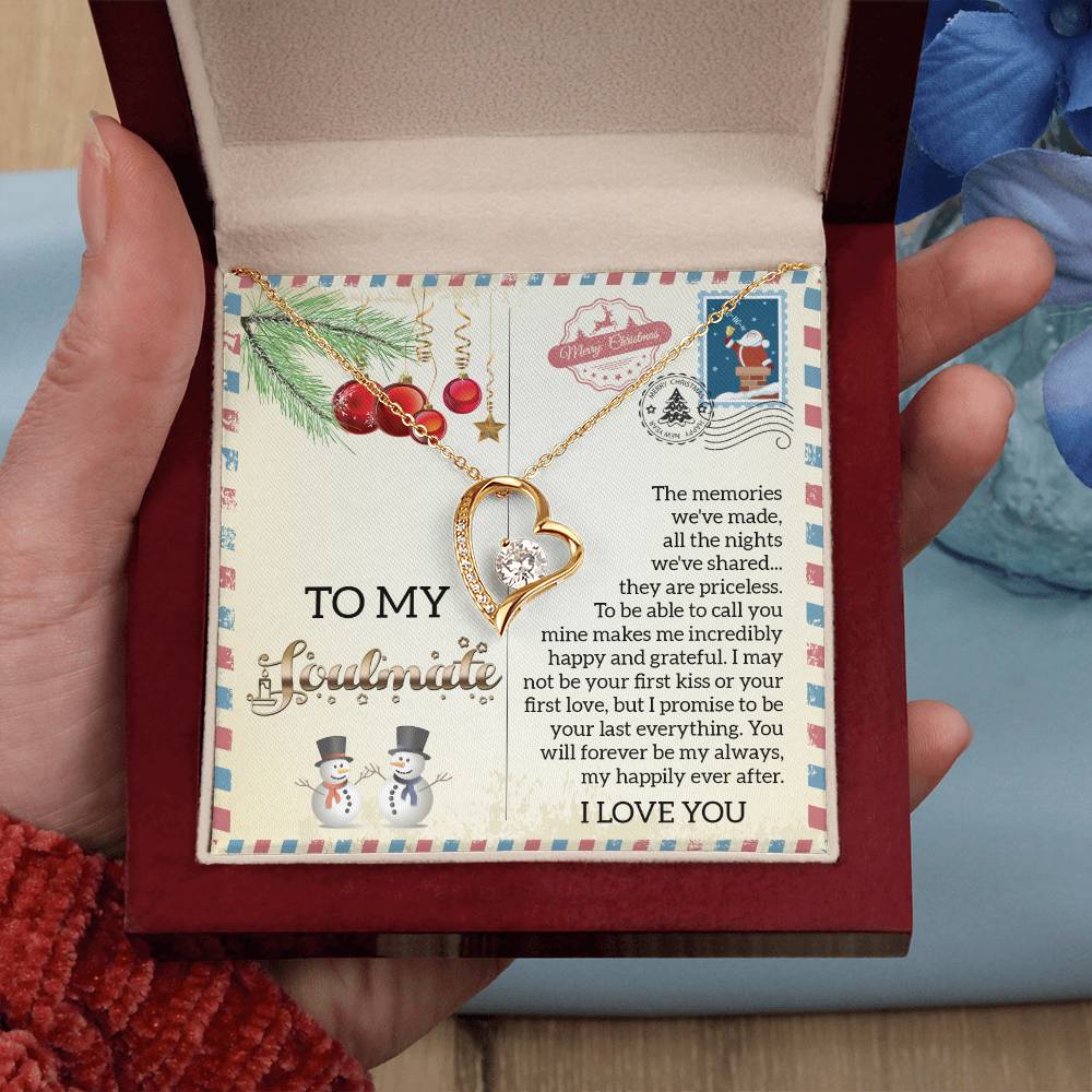 A hand opens a box to reveal the Soulmate-Last Everything - Forever Love Necklace, featuring a gold heart-shaped pendant adorned with a sparkling cubic zirconia. Inside the box is a card addressed "To my soulmate," embellished with snowman illustrations and an affectionate message of love and gratitude.