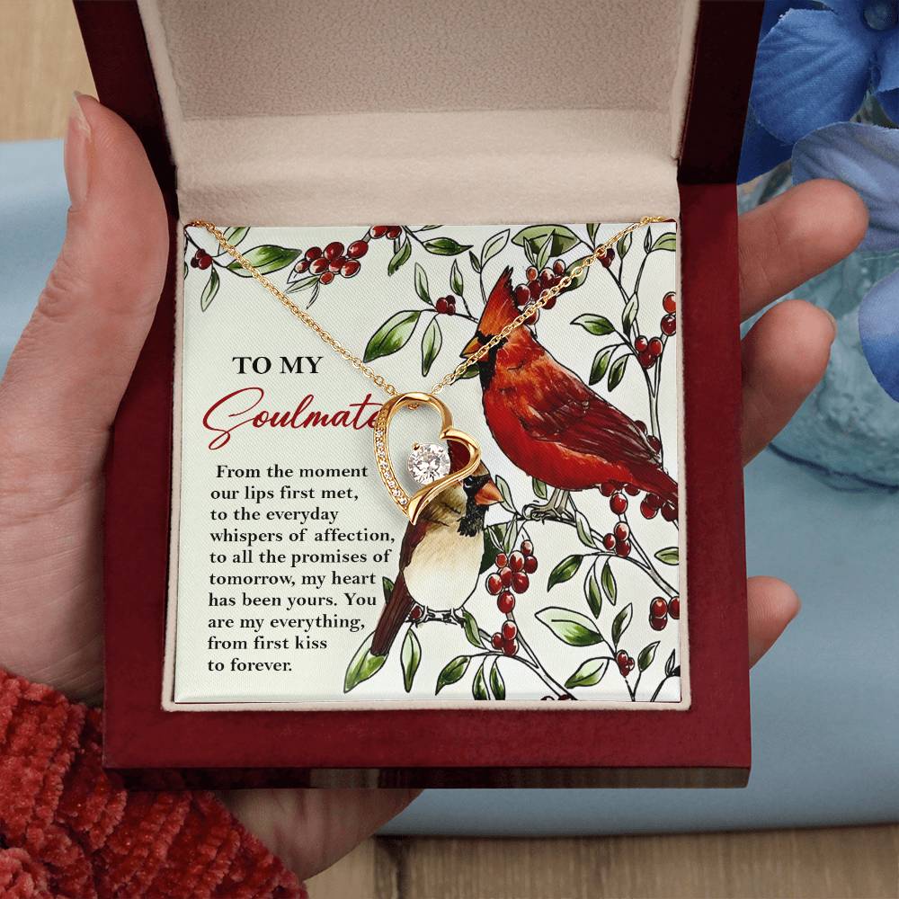 A hand holds an open jewelry box revealing a Soulmate-First Kiss - Forever Love Necklace with a heart and CZ crystal. Behind the pendant is a card adorned with a cardinal illustration and the title "To My Soulmate.