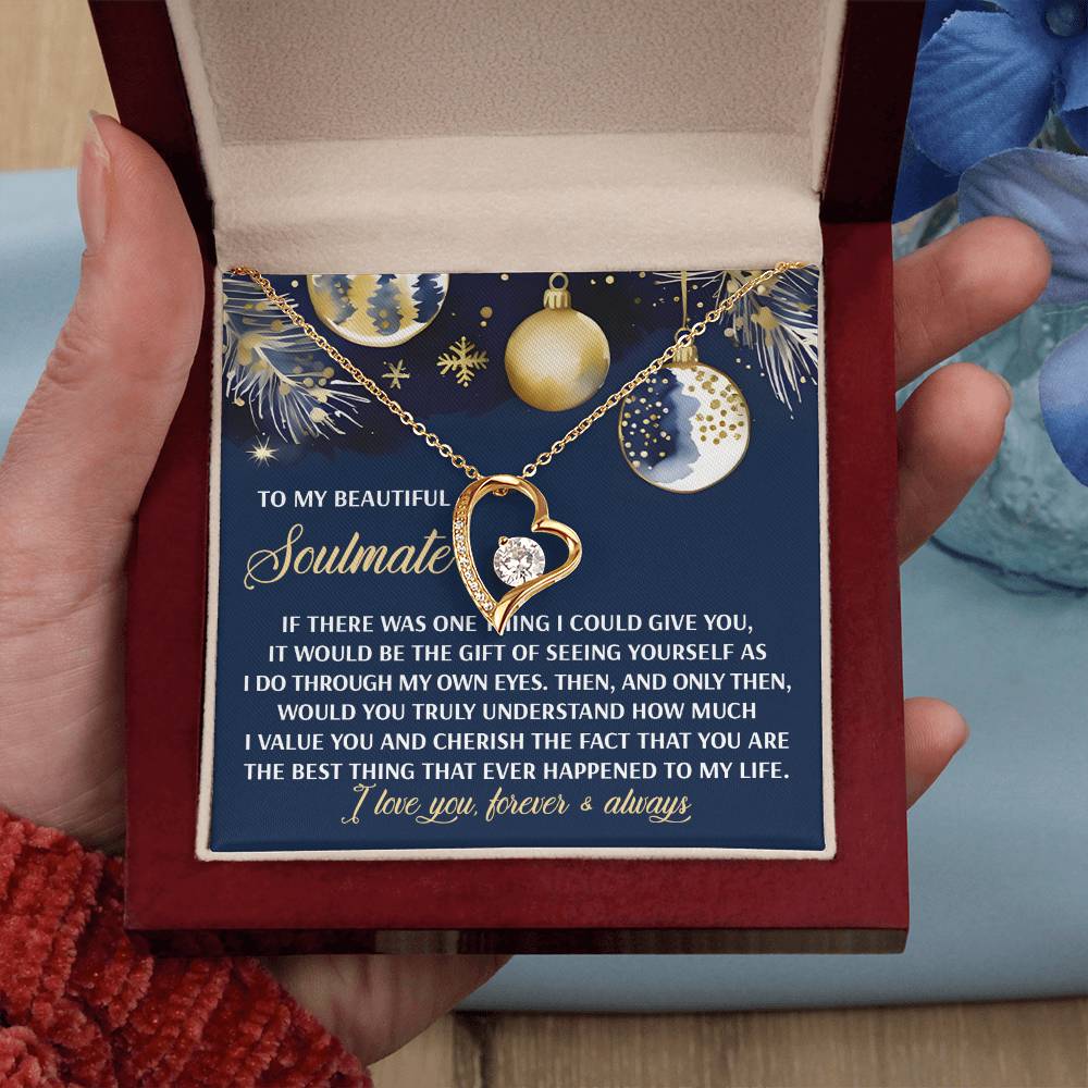 A hand holds an open jewelry box, unveiling a Soulmate-One Thing - Forever Love Necklace with a gold finish and a heart-shaped pendant that sparkles like cubic zirconia. An accompanying festive card, adorned with ornaments and snowflakes, carries a heartfelt message about love.