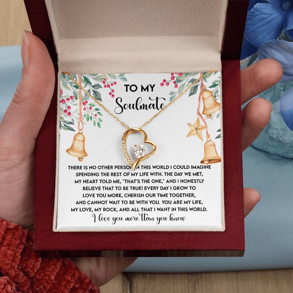 A hand holding a box unveils the Soulmate-Be With You - Forever Love Necklace, featuring a gold finish, accompanied by a heartfelt message. The box is decorated with floral designs and small bell illustrations, lending an elegant touch to the precious cubic zirconia necklace inside.