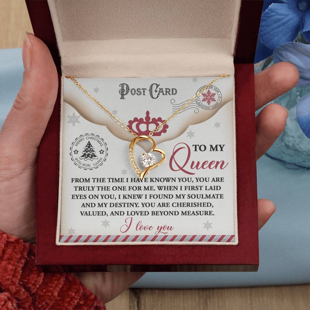 A hand presents a red jewelry box containing the Soulmate-The One - Forever Love Necklace, showcasing a heart-shaped pendant with a white gold finish and CZ crystal. A postcard message addressed "To My Queen" accompanies the gift, while nearby blue flowers enhance the romantic setting.