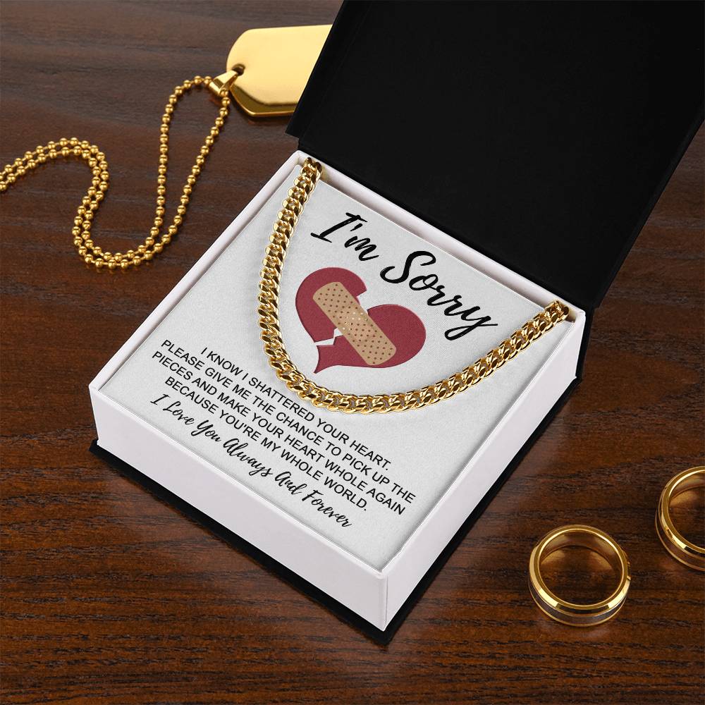 The Sorry-The Pieces - Cuban Link Chain in sleek gold rests in a black box with a card that reads "I'm Sorry" and an apology message below it. Nearby, two 14K yellow gold rings and a polished stainless steel pendant lie on the wooden surface next to the box.