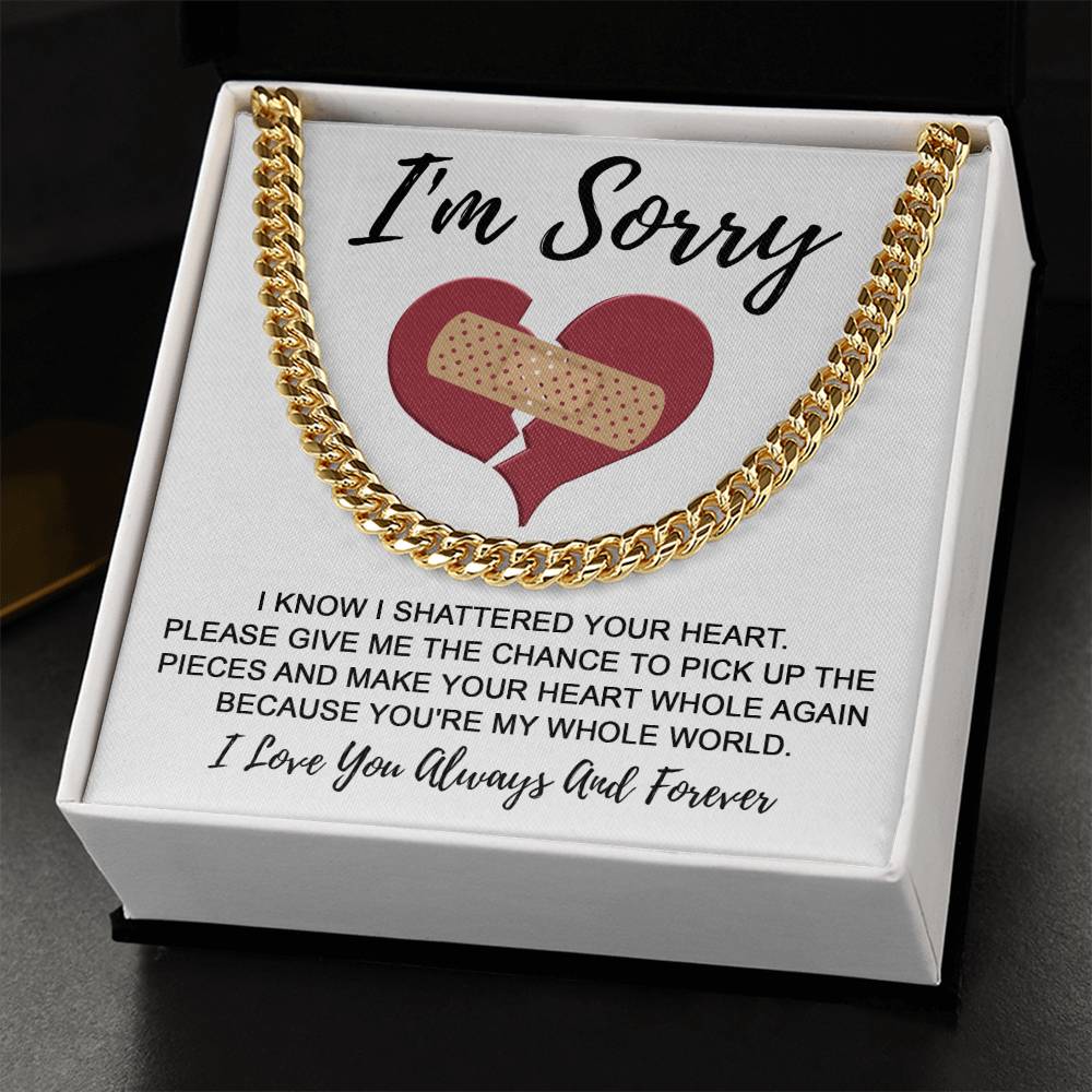 A 14K yellow gold Cuban Link Chain necklace, named "Sorry-The Pieces - Cuban Link Chain," rests on a white cushion inside a box. The box's lid features a heart with a bandage and reads: "I’m sorry. I know I shattered your heart… because you’re my whole world. I love you always and forever.