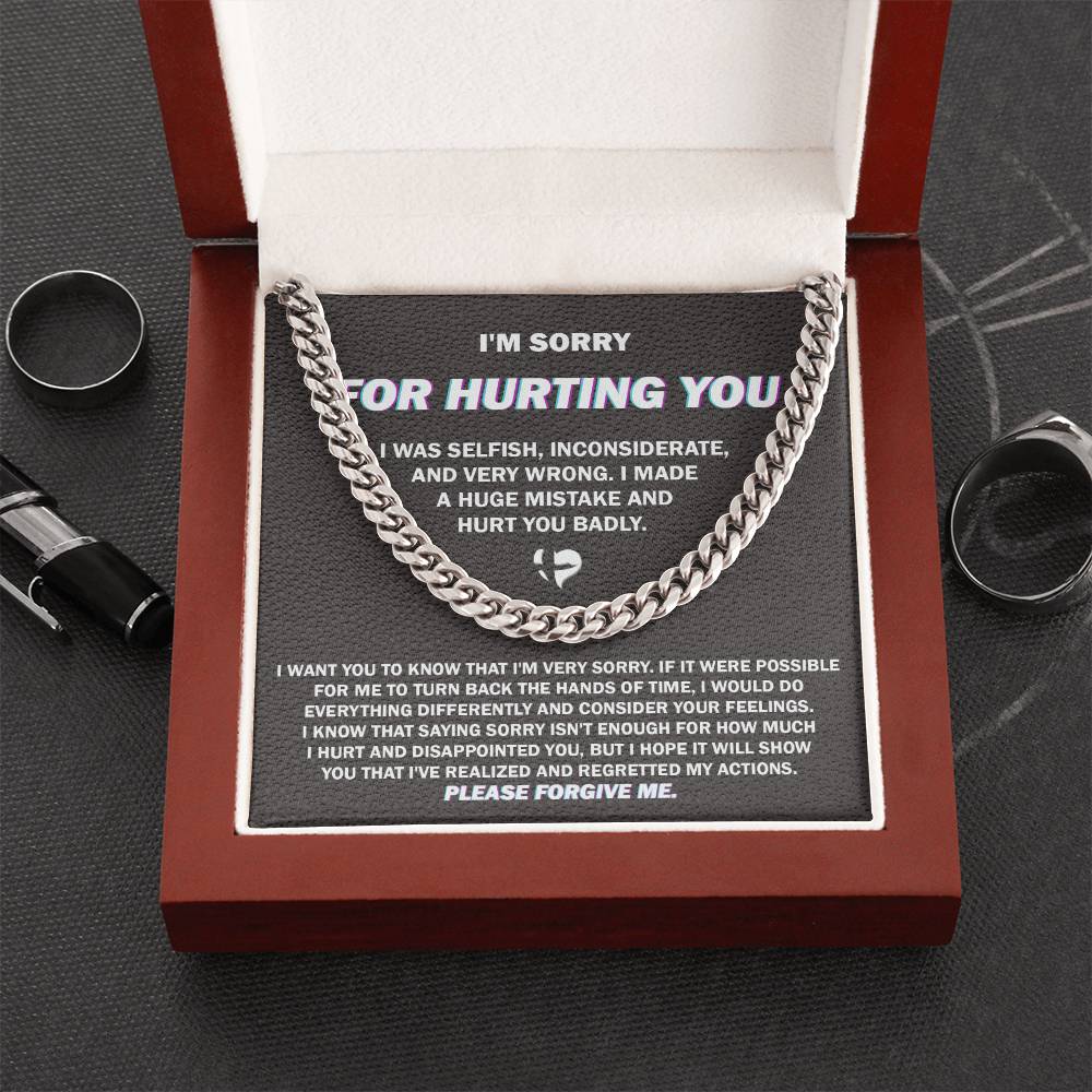 A polished stainless steel Cuban link chain, from the "Sorry, Hurt You Badly" collection, lies inside an open wooden box. The box's lid displays a message that reads, "I'm sorry for hurting you. I was selfish, inconsiderate, and very wrong. I made a huge mistake and hurt you badly. Please forgive me." Several rings and a pen are scattered around the box.