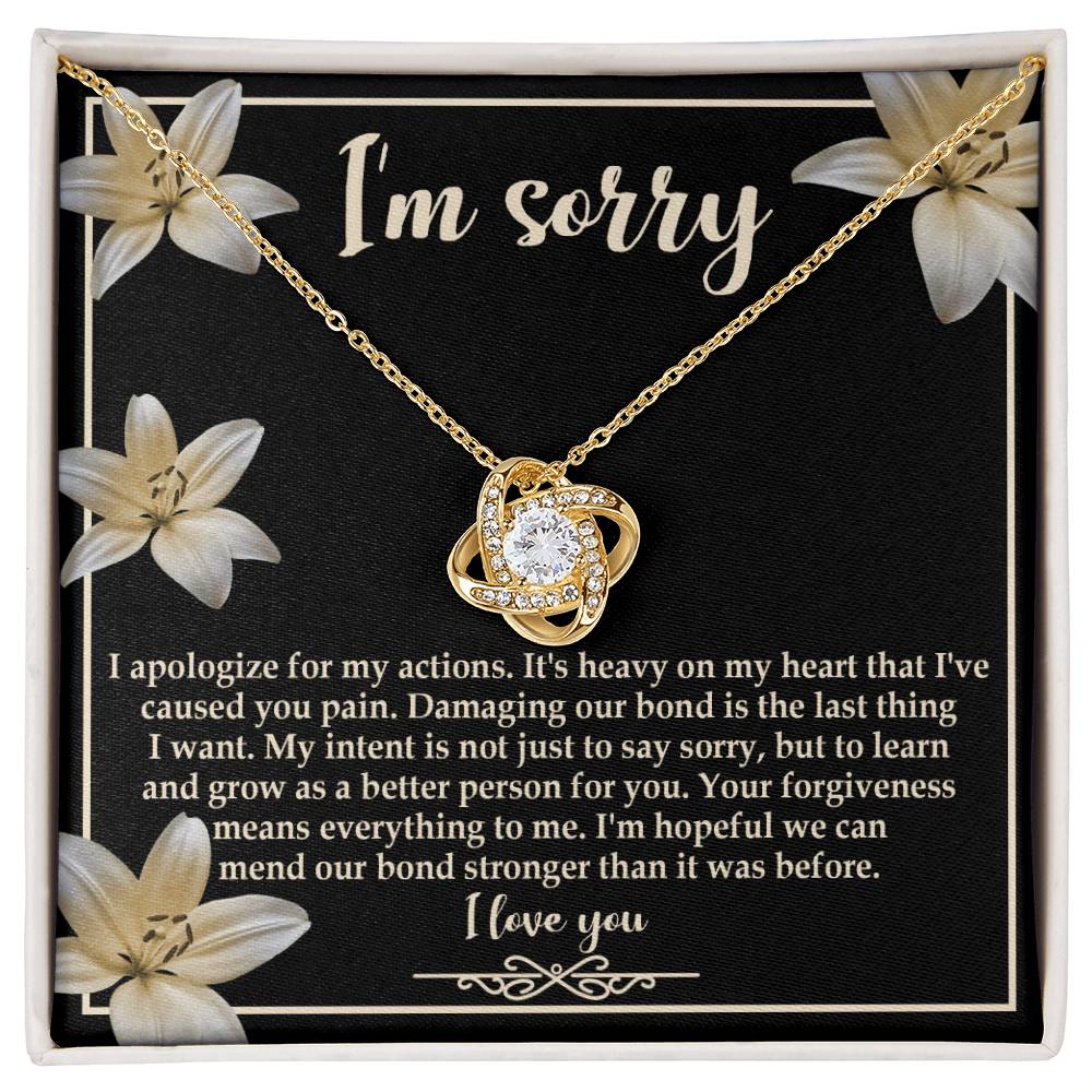 The "Sorry-The Last Thing - Love Knot Necklace" showcases a love knot pendant against a striking black background adorned with white flowers. It's accompanied by an apology message that reads, "I'm sorry. I apologize for my actions... I'm hopeful we can mend our bond stronger," and is beautifully finished with white gold and shimmering cubic zirconia crystals.