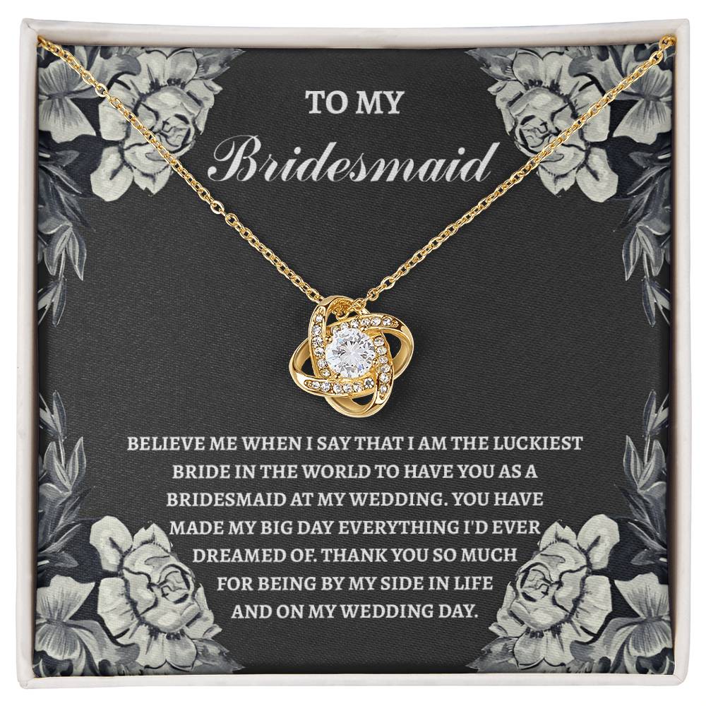 The "To Bridesmaid, The Luckiest Bride - Love Knot Necklace," adorned with cubic zirconia crystals, is elegantly presented on a gift box. The box comes with a heartfelt message expressing gratitude to a bridesmaid. Floral illustrations gracefully decorate the border, adding a charming touch to this piece available in either white gold or yellow gold finish.