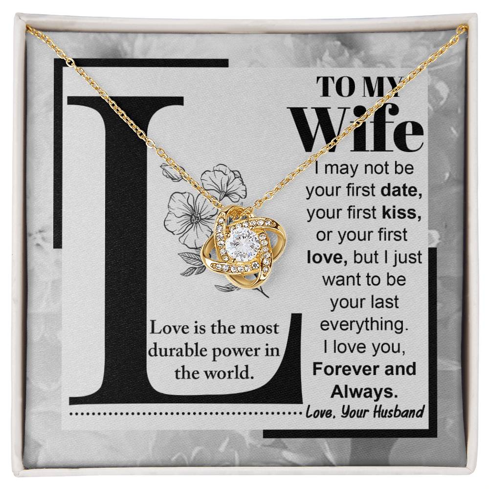 A "to Wife, Love Is - Love Knot Necklace" with a knot design is displayed on a card featuring a printed message to a wife about lasting love, signed from a husband. The piece sparkles with cubic zirconia crystals and boasts an elegant gold finish.