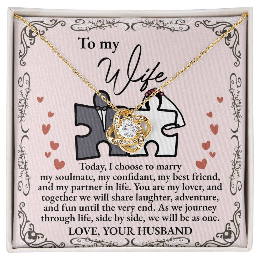 A "To Wife, Be As One - Love Knot Necklace" with intertwined hearts is placed on a card adorned with cubic zirconia crystals, expressing love and commitment from a husband to his wife. The card features a heart and puzzle piece design, available in white gold or yellow gold finish.