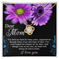 The "To Mom, Held My Hand - Love Knot Necklace" with a crystal pendant sits beautifully in a jewelry box. The box reads: Dear Mom, You held my hand...always be tied to home. I love you. Adorned with cubic zirconia crystals, it's the perfect personalized gift. The background features two purple flowers.