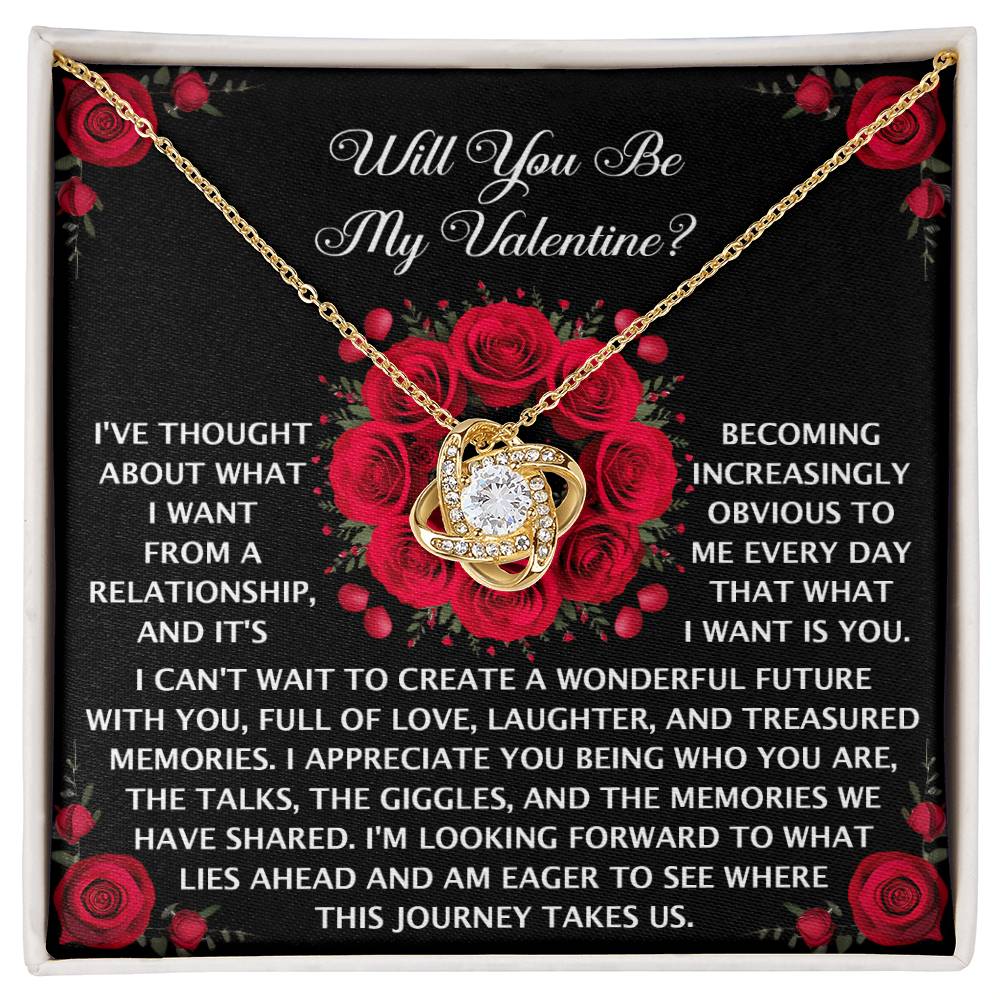 The Soulmate-What I Want - Love Knot Necklace, heart-shaped and adorned with a central Cubic Zirconia gemstone, elegantly glistens on fabric amidst red roses—a romantic message about future relationships softly whispers below.