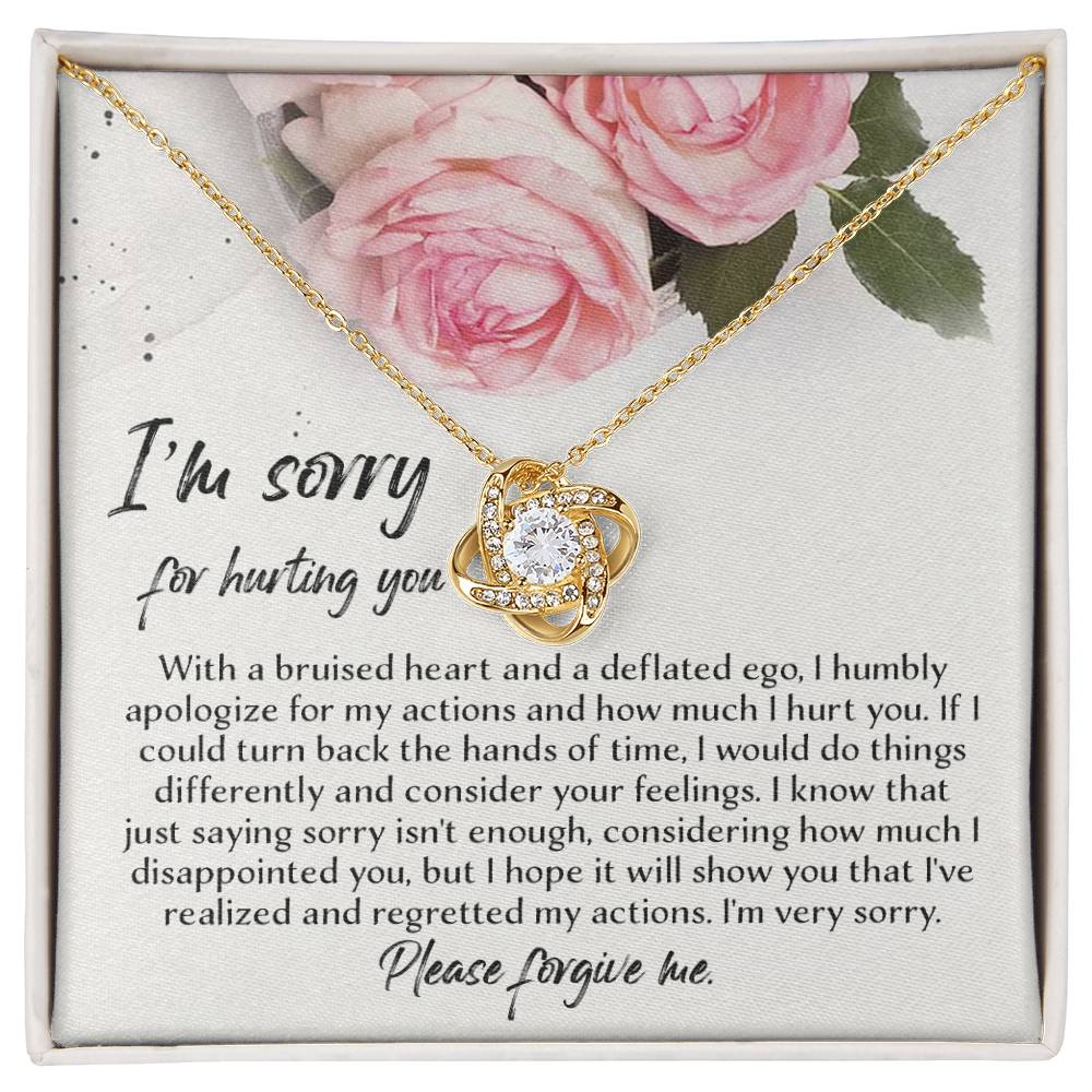 The "Sorry-Regretted My Actions - Love Knot Necklace," crafted in 14k white gold with an intricate pendant adorned with cubic zirconia crystals, is displayed against a backdrop featuring a printed apology note surrounded by roses. The note expresses regret and asks for forgiveness for past actions.