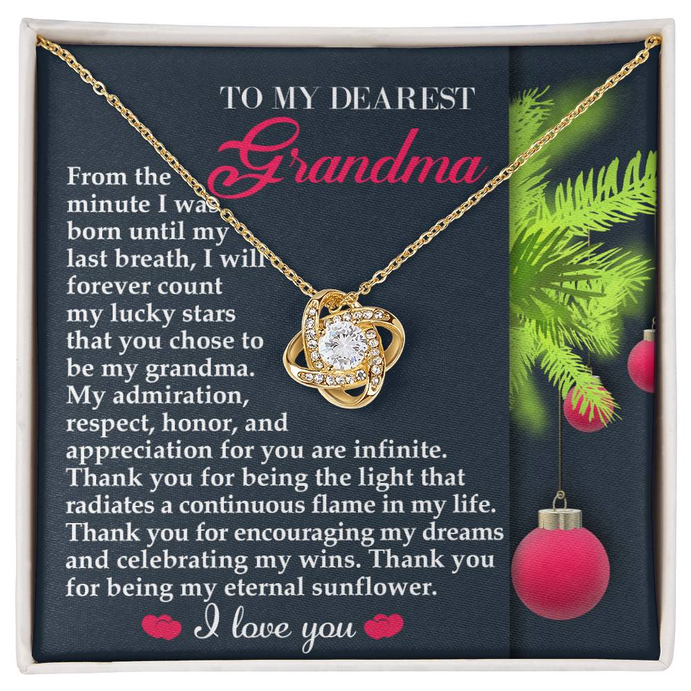 The Grandma-Eternal Sunflower - Love Knot Necklace, showcasing a circular pendant with a white gold finish and sparkling cubic zirconia, is gracefully displayed on a green card. It includes the heartfelt message "To My Dearest Grandma" and "I love you," surrounded by festive holiday decorations.