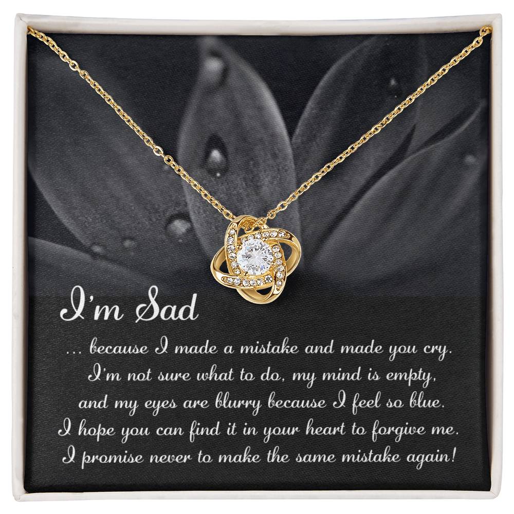 The "Sorry-Feel So Blue - Love Knot Necklace," featuring a stunning knot pendant adorned with cubic zirconia crystals, is displayed over a card with an apologetic message starting with "I'm Sad" and set against a floral background. This personalized gift beautifully combines elegance and heartfelt sentiment.