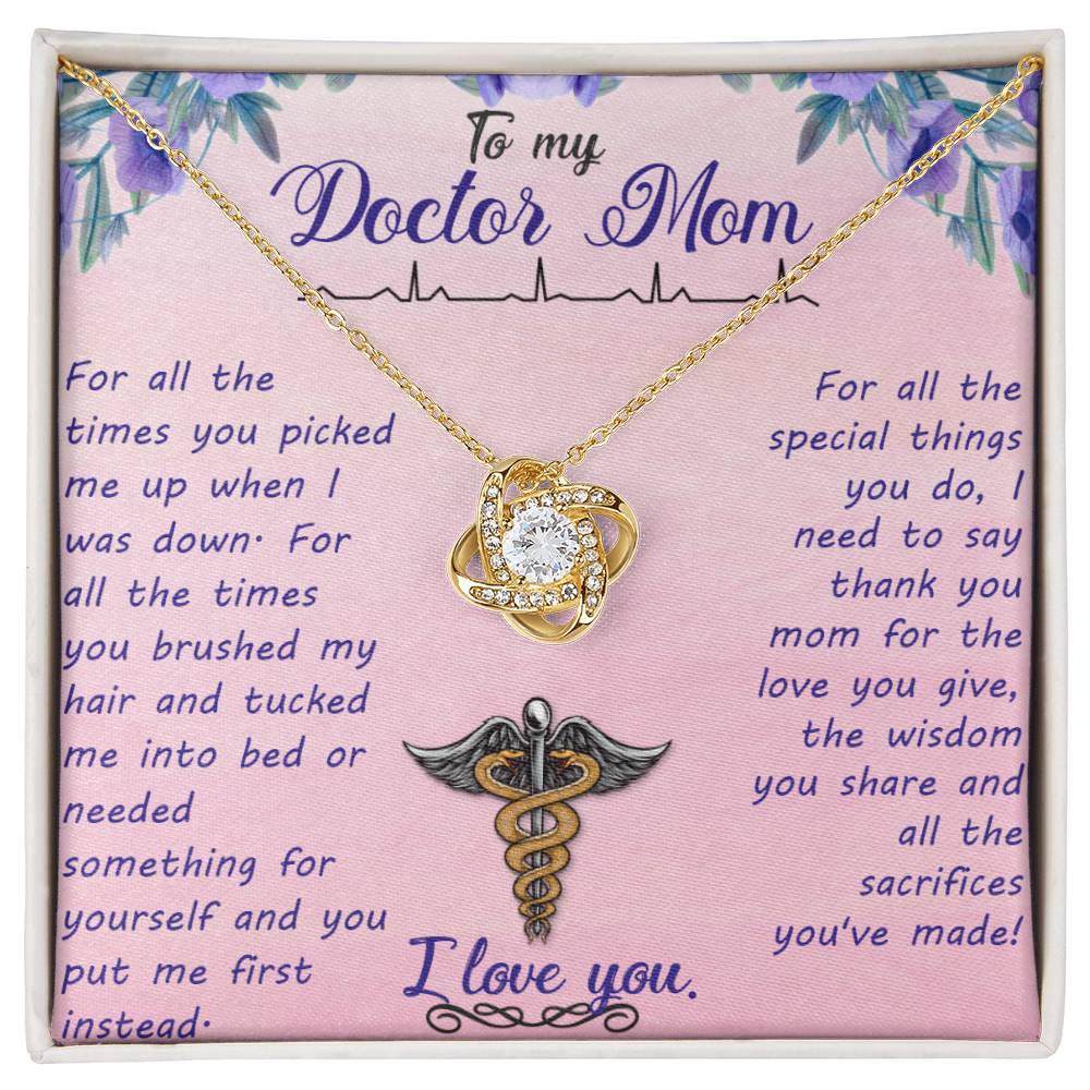 A gold-colored To My Doctor Mom, For All The Times You Picked Me Up - Love Knot Necklace with a heart-shaped pendant, adorned with cubic zirconia crystals and a medical caduceus symbol, presented on a pink card with a heartfelt message to a mother who by ShineOn Fulfillment.