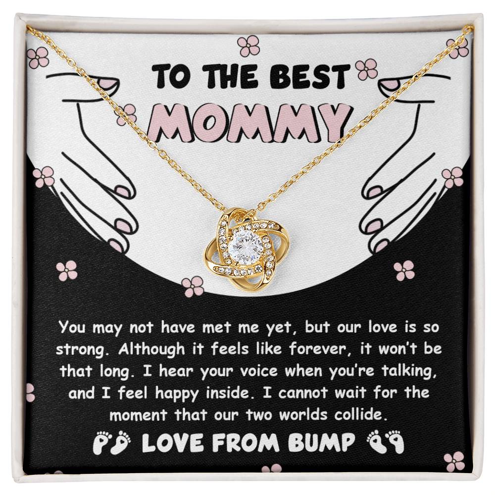 A gold Love Knot Necklace, named "To Mom To Be, Two Worlds Collide," featuring an intertwined heart pendant adorned with cubic zirconia crystals, is enclosed in a box with a message that reads "To the best mommy, love from bump." This heartfelt message beautifully expresses the unbreakable bond and love from an unborn baby.