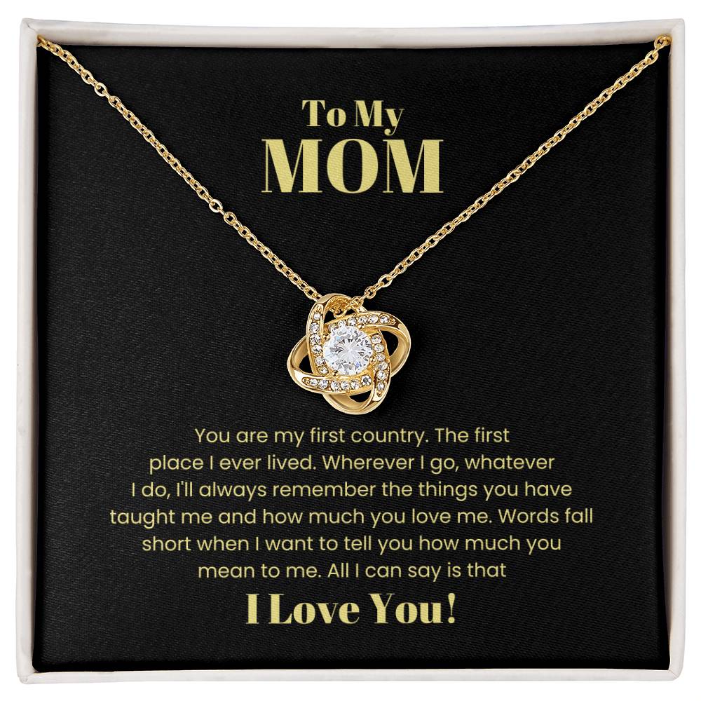 The "To My Mom, You Are My First Country - Love Knot Necklace" in 14k white gold with a diamond pendant is showcased on a black cushion. The engraved message above it reads "To My Mom," accompanied by a heartfelt note ending with "I Love You.