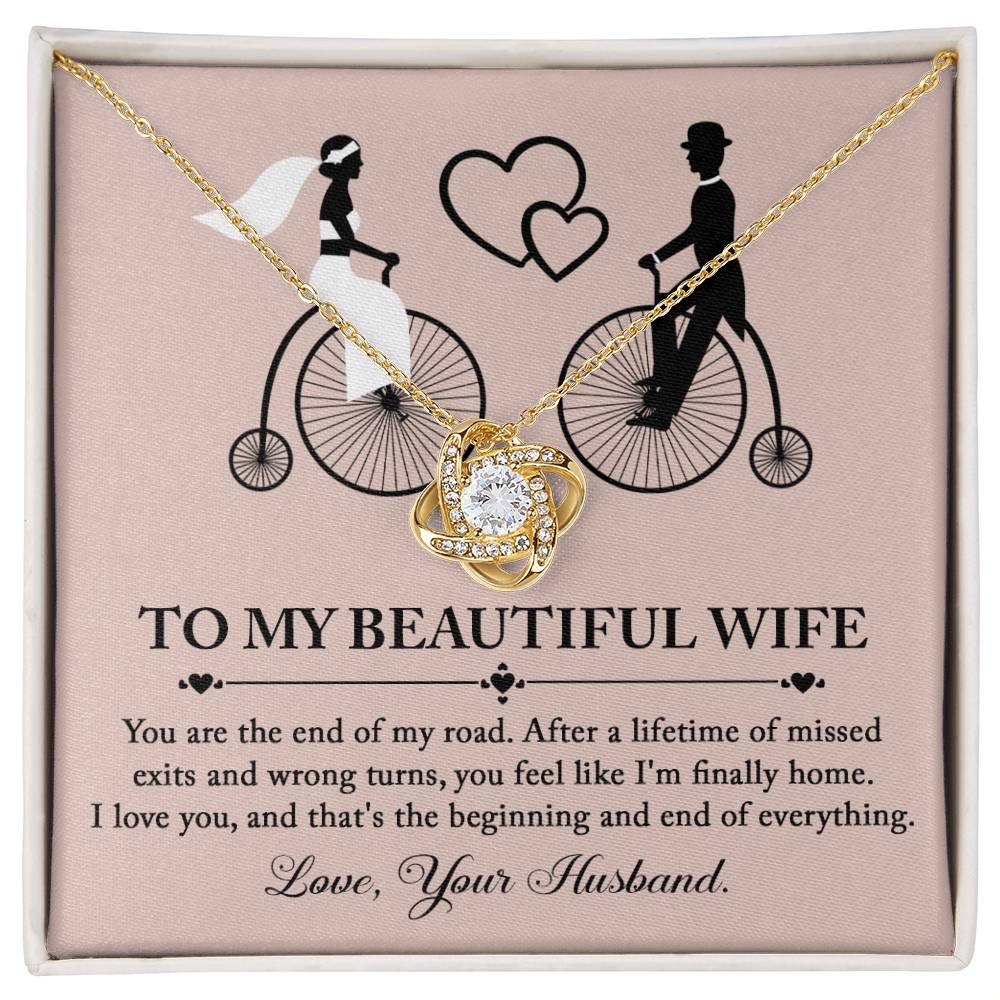 To Wife, I'm Finally Home - Love Knot Necklace with intertwined circle pendant on a card featuring an illustration of a couple on bicycles. The necklace, adorned with cubic zirconia crystals and a stunning gold finish, comes with a card that reads: 'To my beautiful wife' followed by a romantic message and ending with 'Love, Your Husband.'