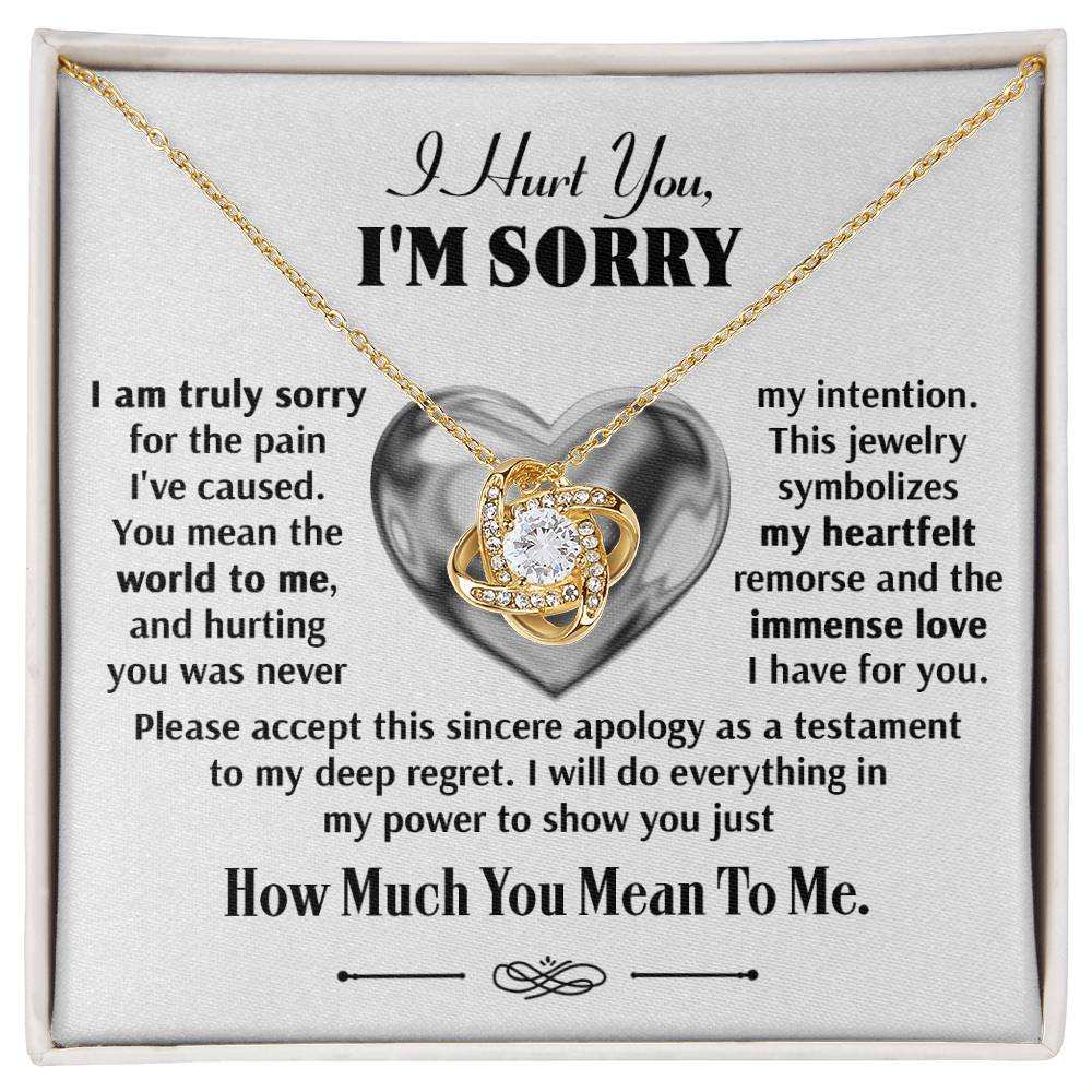 The Sorry-Sincere Apology - Love Knot Necklace, featuring a gold pendant with a cubic zirconia crystal centerpiece, is displayed in an open box. The background contains a heartfelt apology message expressing remorse and dedication to the recipient.