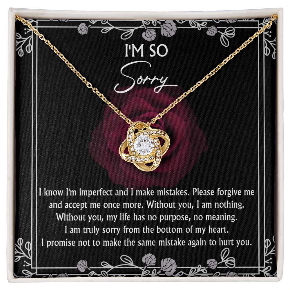 The "Sorry-I Am Nothing - Love Knot Necklace" with a pendant featuring interlocking curves and a central cubic zirconia crystal is displayed in a box. The necklace has a yellow gold finish, and the box's cover shows an apology message decorated with a red rose graphic in the background.