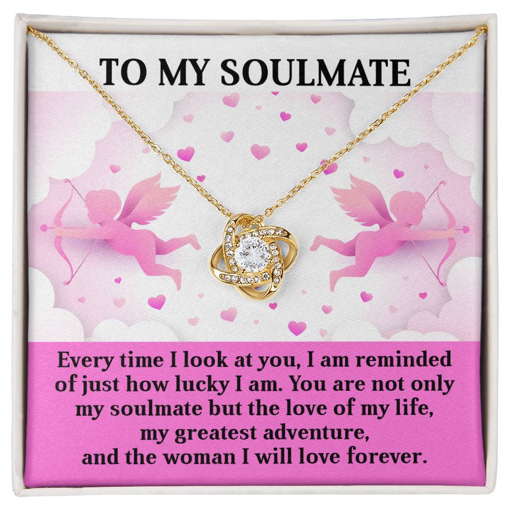 The Soulmate-Greatest Adventure Love Knot Necklace, adorned with cubic zirconia crystals and a heart-shaped pendant, is set against a pink background with cupid motifs and a soulmate message. It makes for an ideal personalized gift for your love's special occasion.