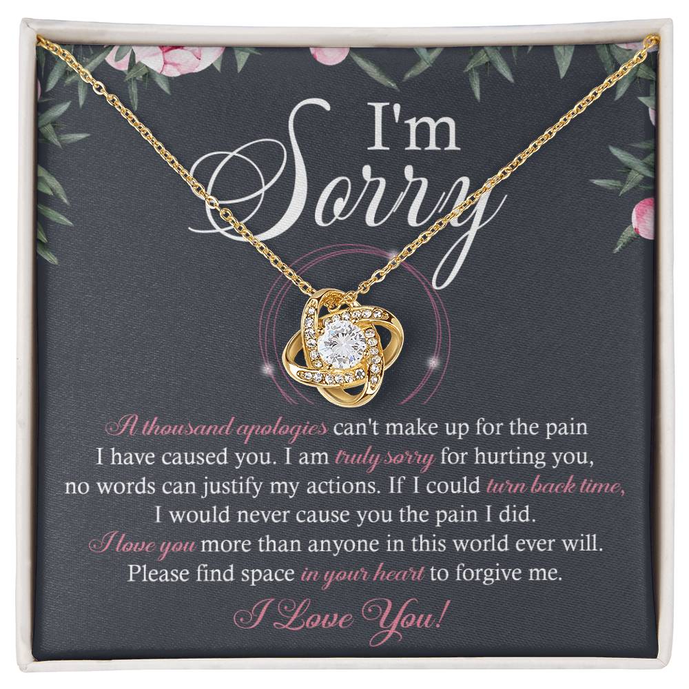 A box containing the "Sorry-World Ever Will - Love Knot Necklace," featuring a central pendant adorned with cubic zirconia crystals. The box lid displays text apologizing and seeking forgiveness, framed by a decorative floral border.