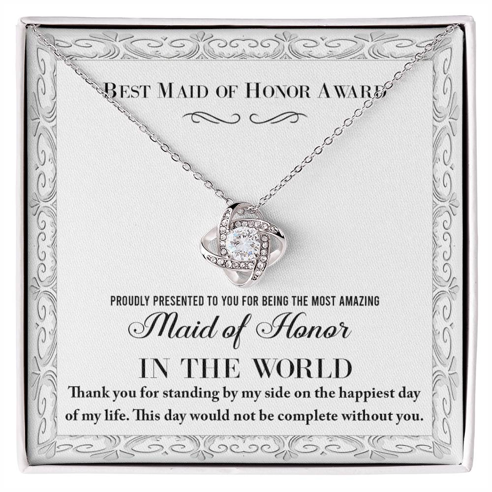 A "To Maid of Honor, Of My Life - Love Knot Necklace," crafted in 14k white gold with a heart-shaped pendant adorned with cubic zirconia crystals, sits elegantly in a jewelry box. The text reads, "Best Maid of Honor Award. Proudly presented to you for being the most amazing Maid of Honor in the world.