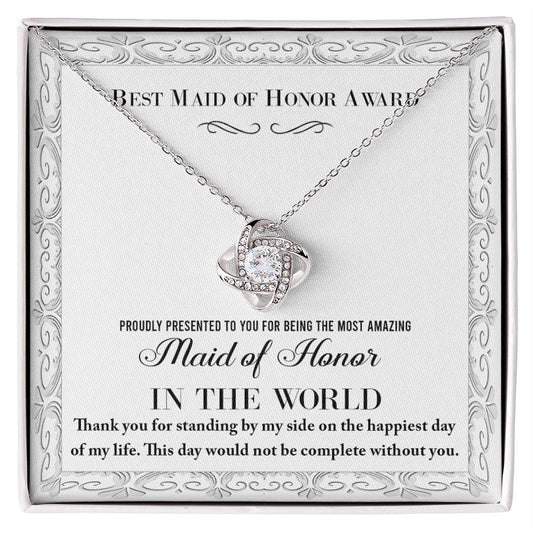 A "To Maid of Honor, Of My Life - Love Knot Necklace," crafted in 14k white gold with a heart-shaped pendant adorned with cubic zirconia crystals, sits elegantly in a jewelry box. The text reads, "Best Maid of Honor Award. Proudly presented to you for being the most amazing Maid of Honor in the world.