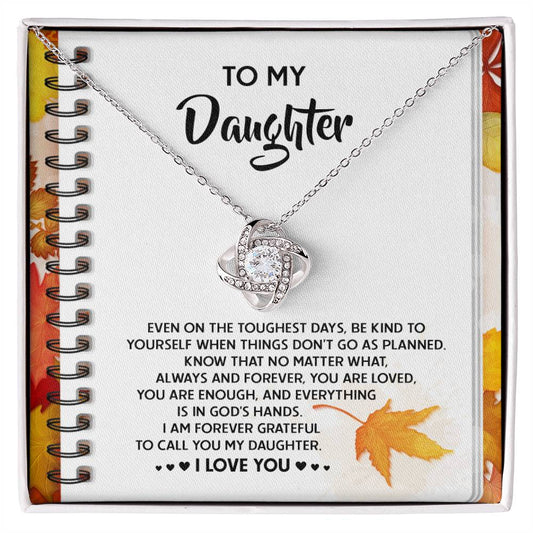 The Daughter-In God's Hands - Love Knot Necklace features a heart-shaped pendant adorned with small cubic zirconia crystals and comes displayed on a message card that reads, "To my Daughter. Even on the toughest days, be kind to yourself...". This personalized gift captures the essence of love and encouragement.