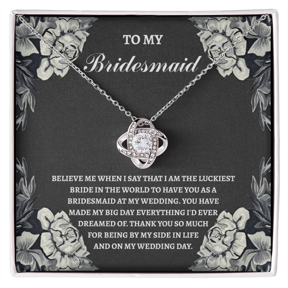 The "To Bridesmaid, The Luckiest Bride - Love Knot Necklace," adorned with cubic zirconia crystals, is elegantly presented on a gift box. The box comes with a heartfelt message expressing gratitude to a bridesmaid. Floral illustrations gracefully decorate the border, adding a charming touch to this piece available in either white gold or yellow gold finish.