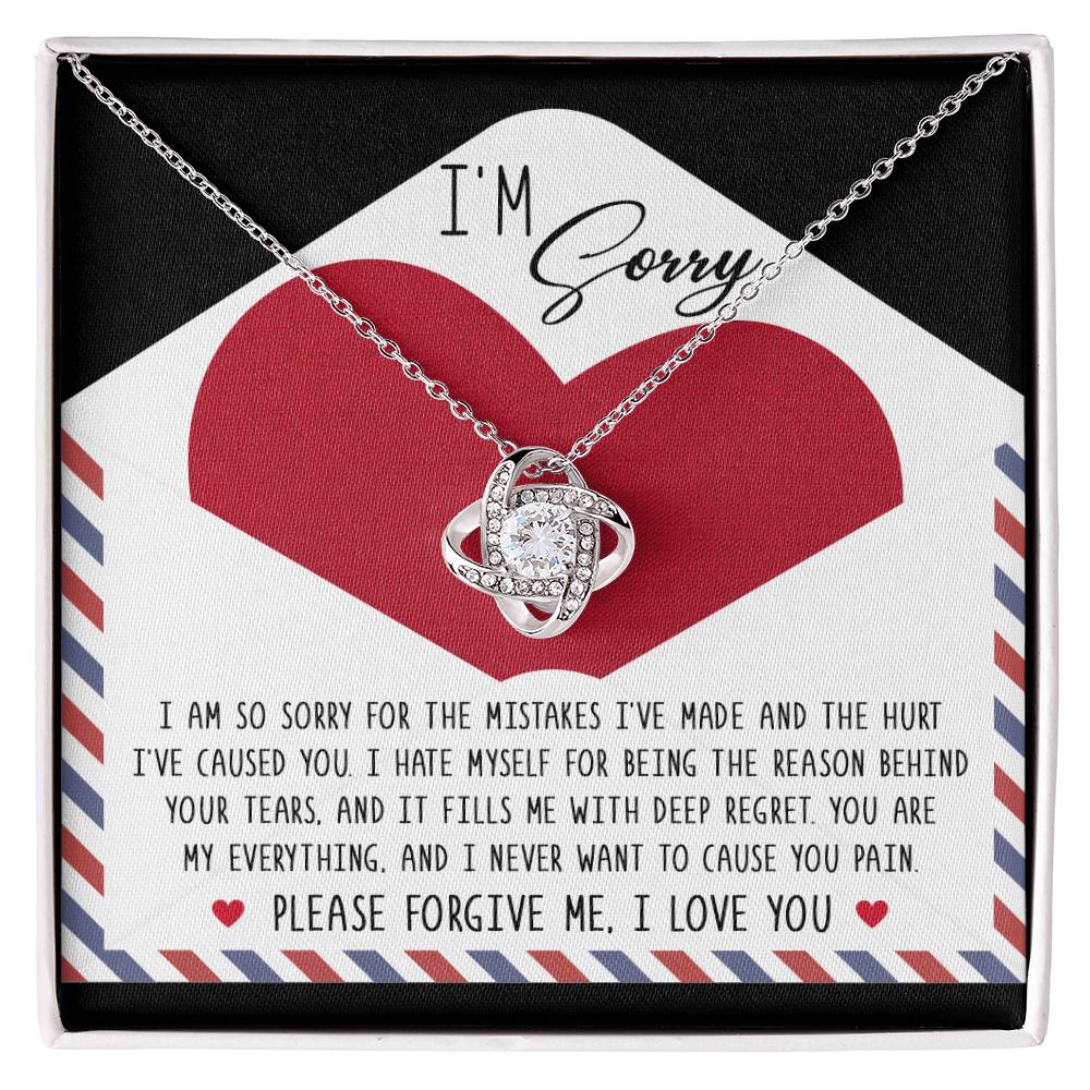 The Sorry-Behind Your Tears - Love Knot Necklace, featuring a silver pendant adorned with cubic zirconia crystals, is presented in a box along with a heartfelt note that reads "I'm Sorry" and an apology message. The background includes a large red heart.