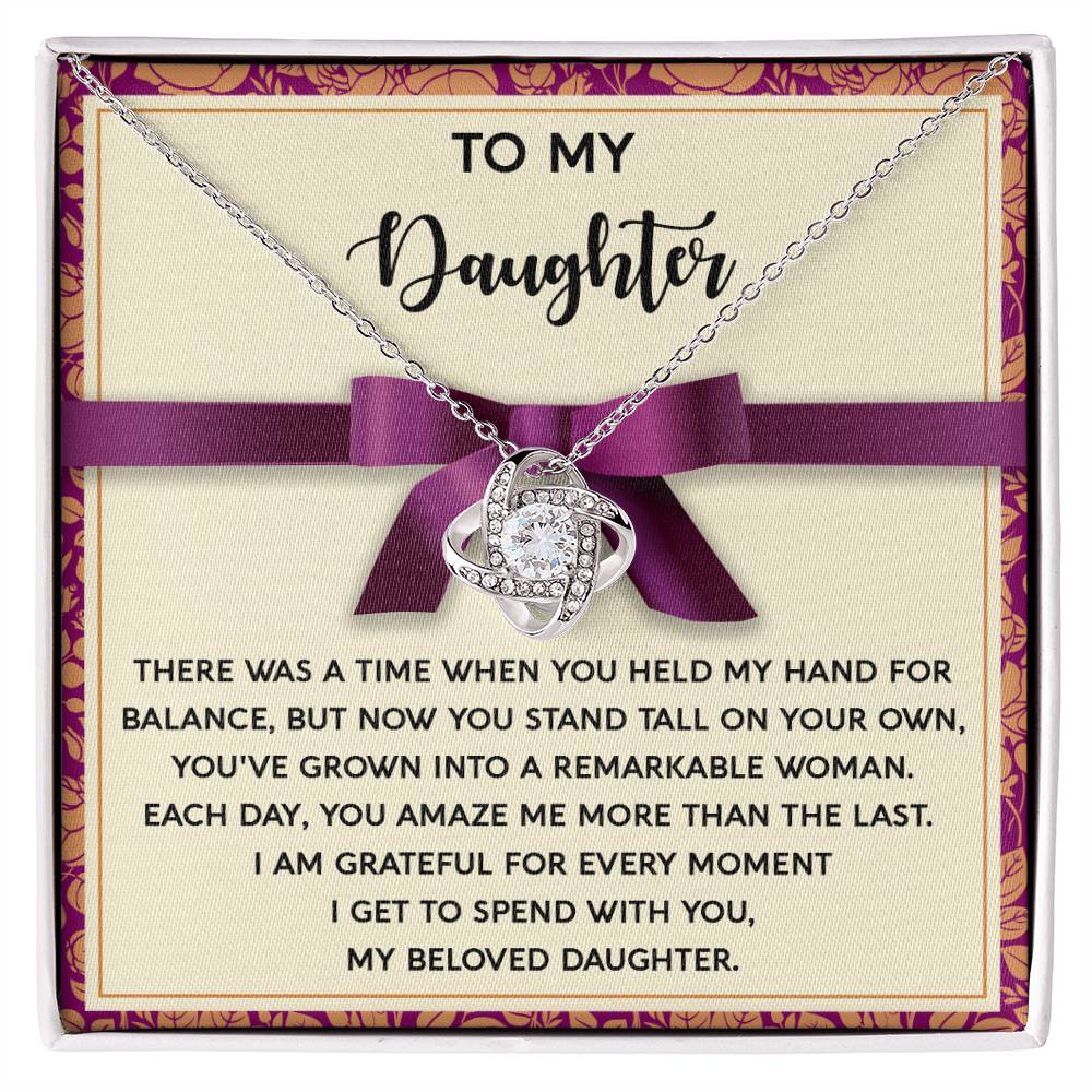 The Daughter-Spend With You - Love Knot Necklace, featuring an elegant intertwined heart design, is presented on a card with a heartfelt message to a daughter, expressing pride and love for her growth. The card, decorated with a purple ribbon and border, beautifully highlights the necklace adorned with cubic zirconia crystals.