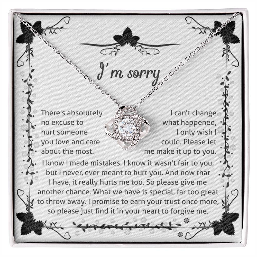 The Sorry-No Excuse - Love Knot Necklace, with a white gold finish, showcases a delicate knot design on a chain adorned with cubic zirconia crystals. The pendant is presented on a box featuring an "I'm sorry" message that expresses regret and seeks forgiveness, framed by a charming floral border at the top.