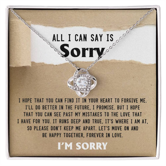 The Sorry-Keep Me Apart - Love Knot Necklace features a sparkling pendant adorned with cubic zirconia crystals, presented in an elegant box that reads, "ALL I CAN SAY IS Sorry" with an apology message below, concluding with "I'M SORRY." It is available in a choice of white gold or yellow gold finish.