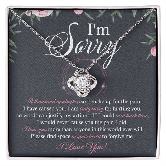 A box containing the "Sorry-World Ever Will - Love Knot Necklace," featuring a central pendant adorned with cubic zirconia crystals. The box lid displays text apologizing and seeking forgiveness, framed by a decorative floral border.