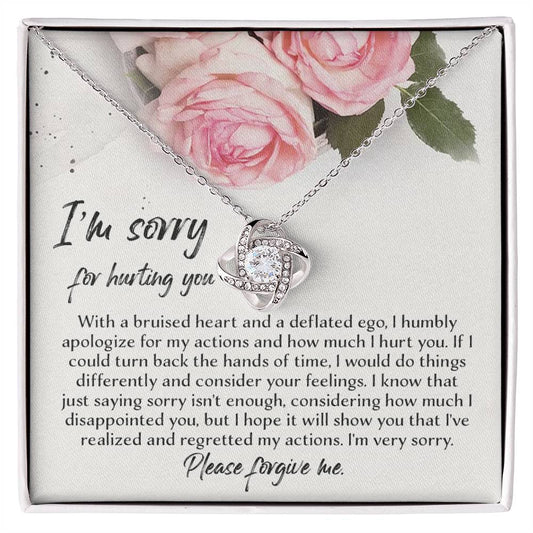 The "Sorry-Regretted My Actions - Love Knot Necklace," crafted in 14k white gold with an intricate pendant adorned with cubic zirconia crystals, is displayed against a backdrop featuring a printed apology note surrounded by roses. The note expresses regret and asks for forgiveness for past actions.