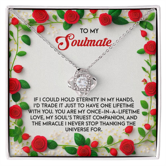 The Soulmate-One Lifetime - Love Knot Necklace, in white or yellow gold finish, is elegantly presented in a jewelry box with cubic zirconia crystals. It includes a heartfelt message about love and soulmates, bordered by red roses and lush green leaves.