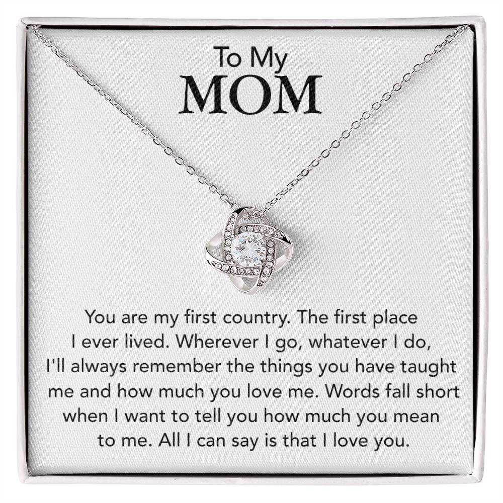 A To My Mom, You Are My First Country - Love Knot Necklace with a heart pendant, adorned with cubic zirconia crystals, is displayed on a box with a personalized heartfelt message to a mother expressing love and gratitude from ShineOn Fulfillment.