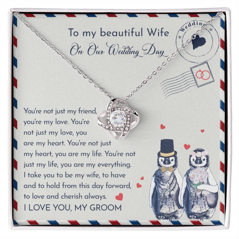 Close-up of a To Wife Wedding, Love And Cherish - Love Knot Necklace with cubic zirconia crystals on a card. The 14k white gold pendant is heart-shaped, and the card has a message for a wife on the wedding day. Two penguins, dressed as a bride and groom, are illustrated at the bottom.